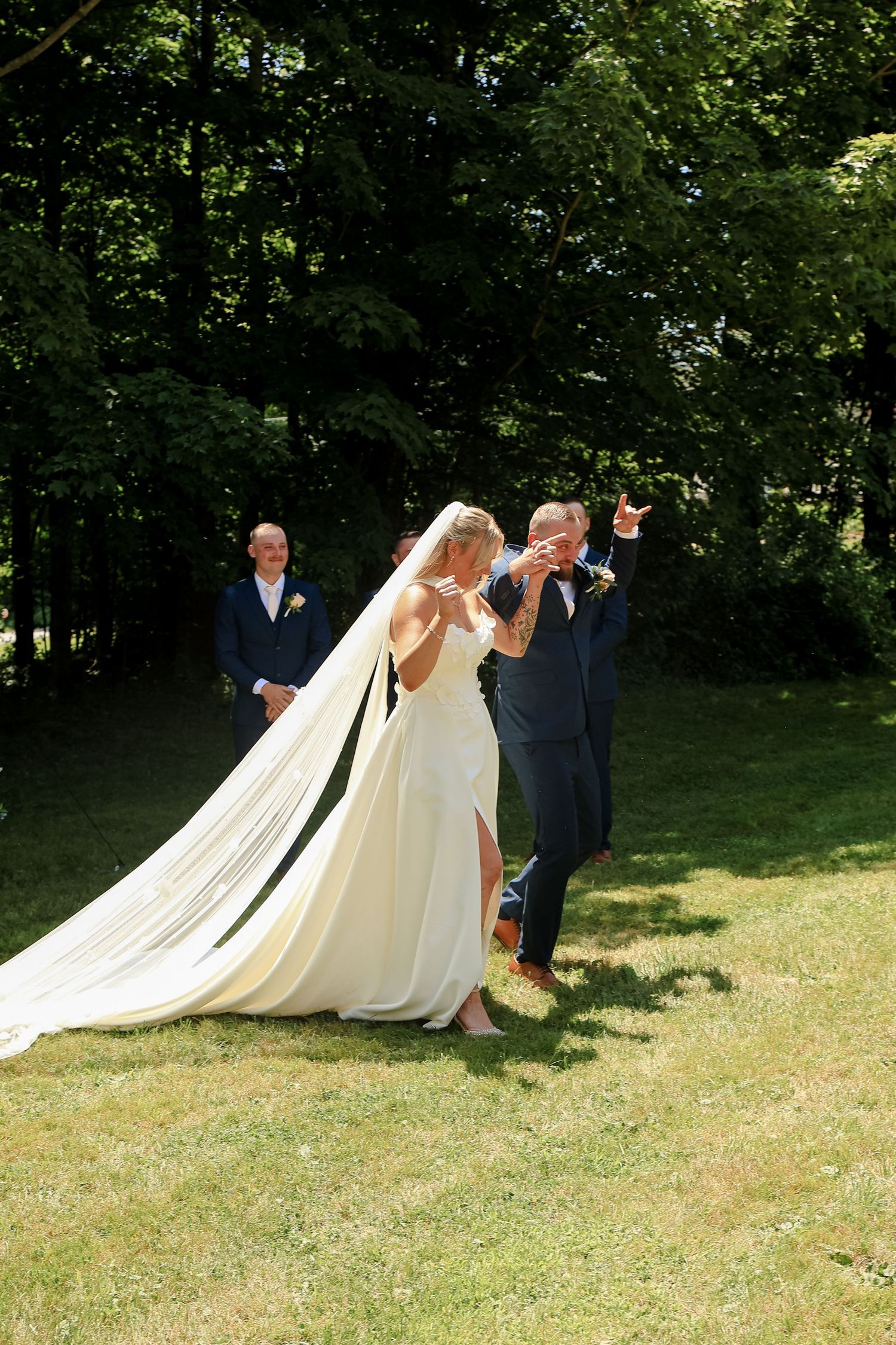 Bride in white gown with long veil celebrates with groom outdoors on sunny lawn surrounded by lush green trees.
