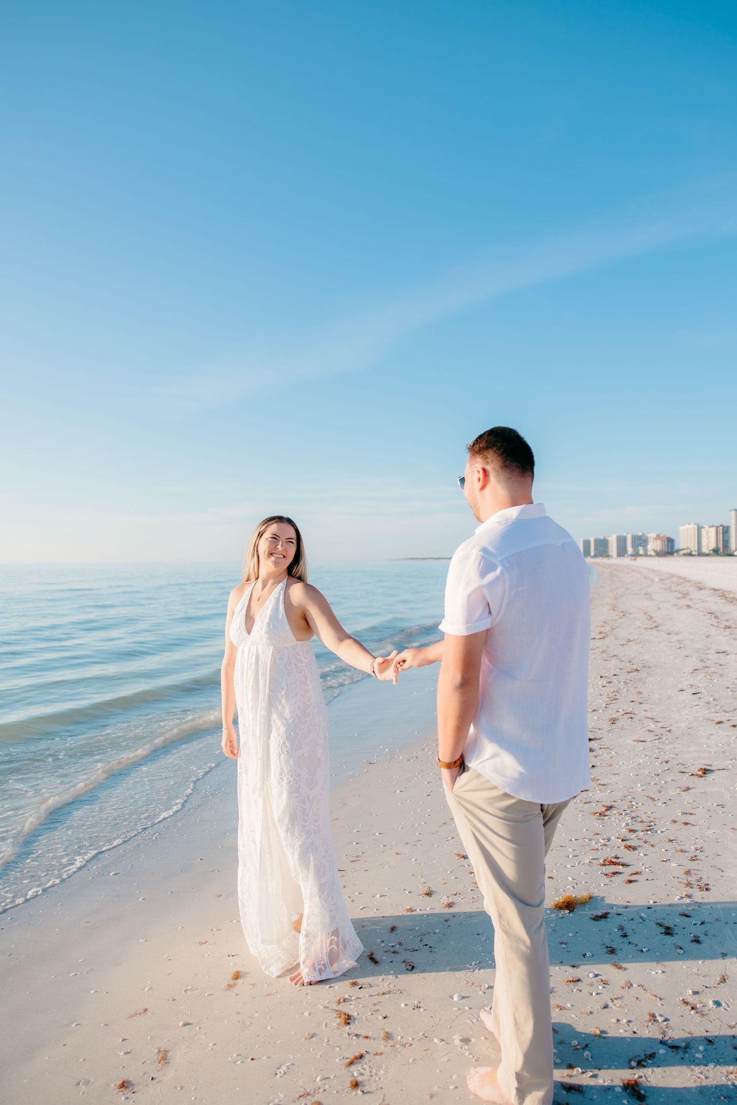 A couple in white outfits walks along a pristine beach with turquoise waters and clear blue skies on a sunny day.