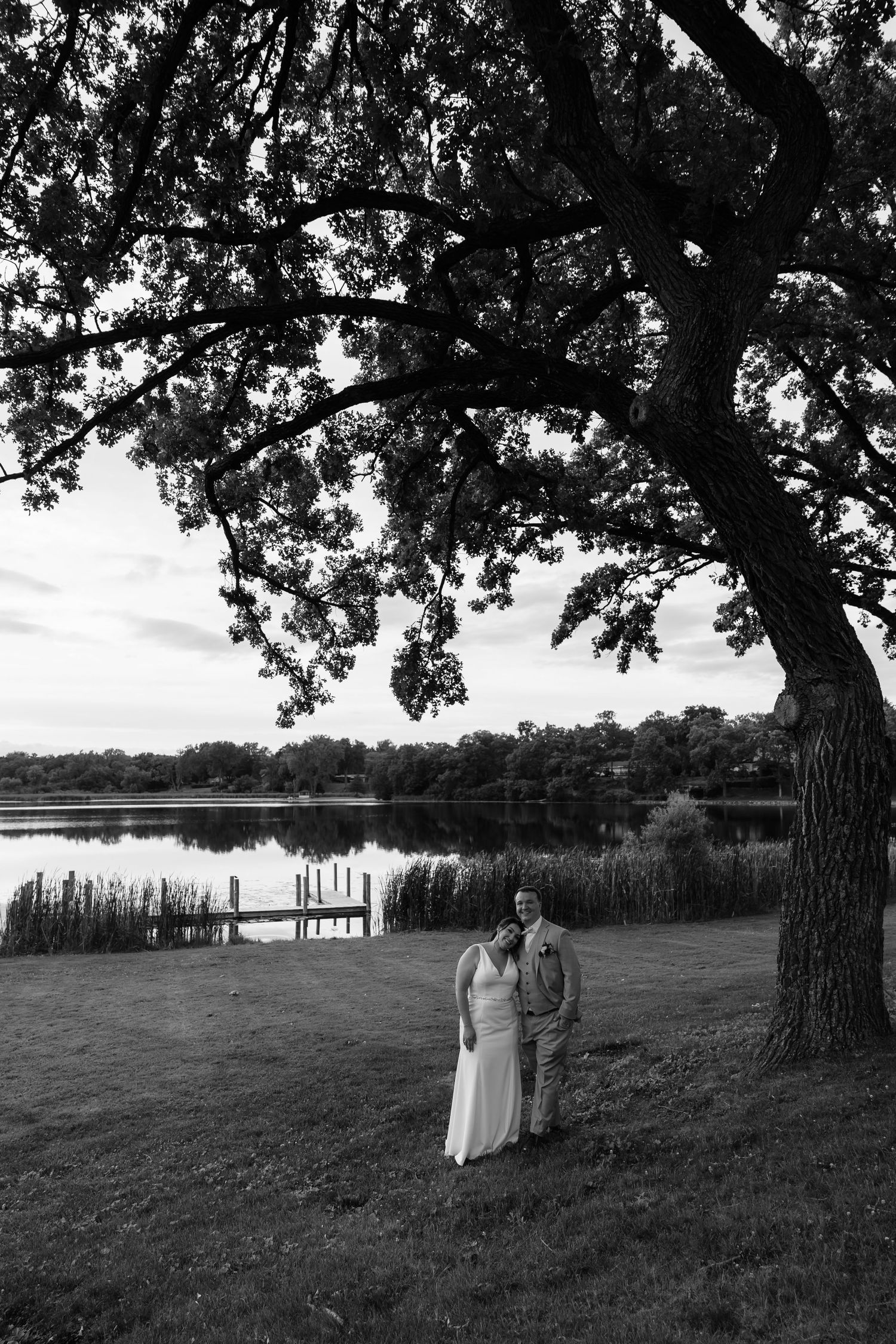 Black and white photo of a wedding couple standing under a large oak tree by a serene lakeside dock.