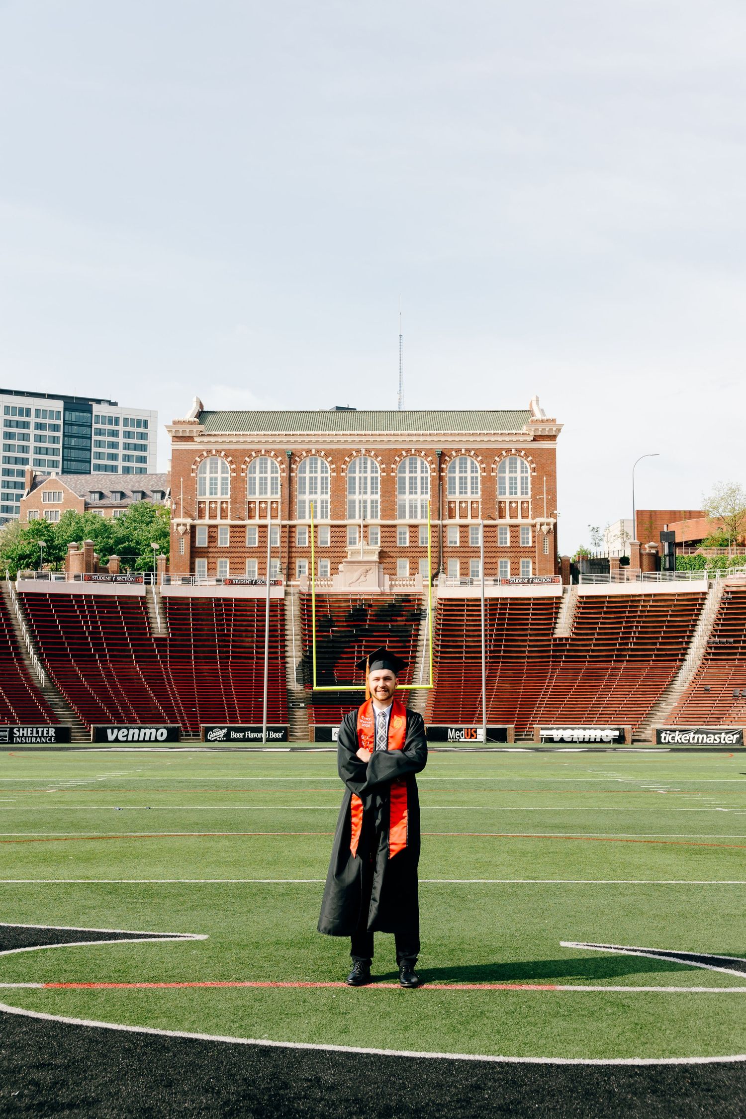 Graduate in cap and gown stands on football field with brick stadium and city buildings in background.