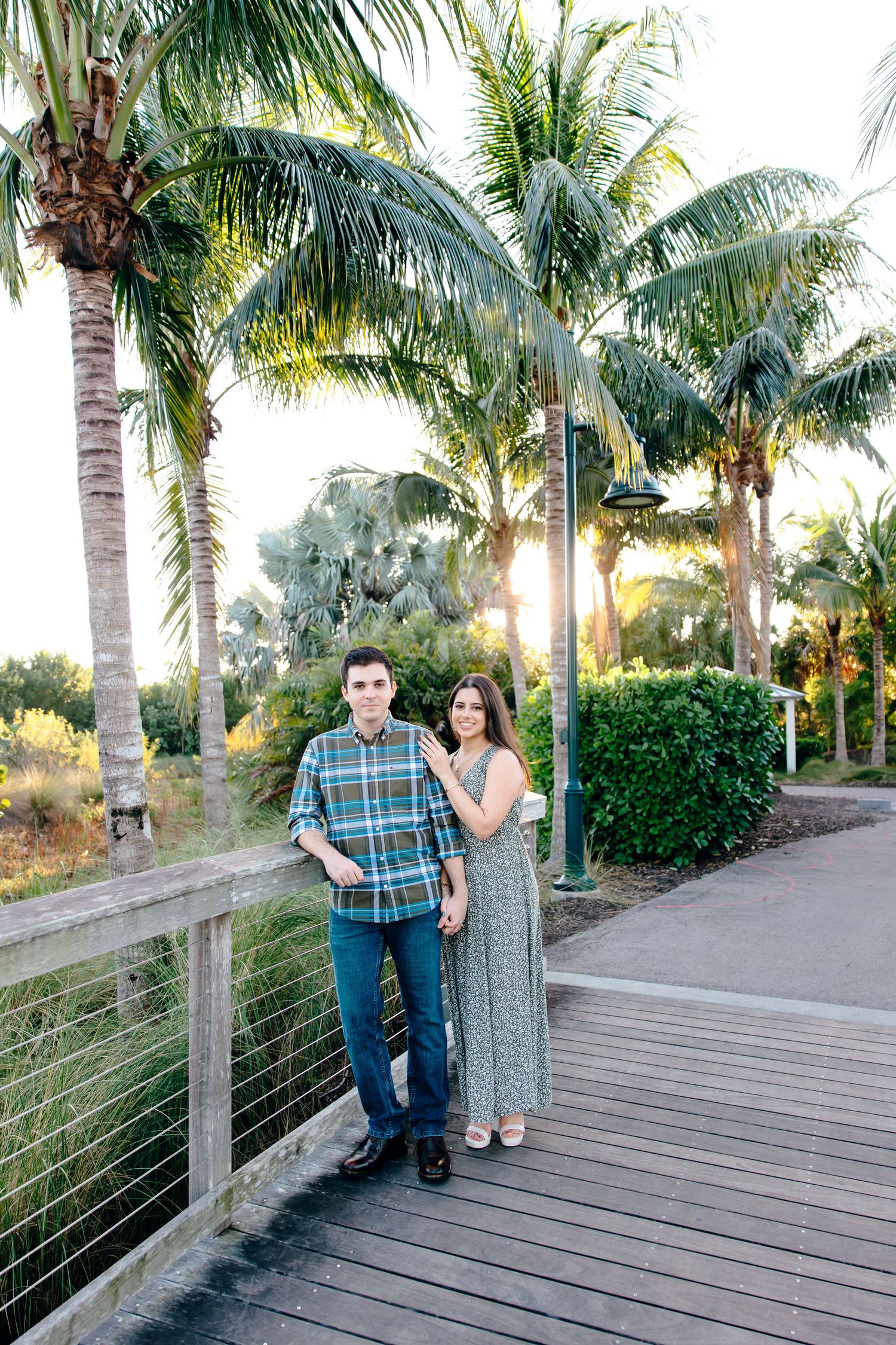 Couple posing on a wooden boardwalk surrounded by lush palm trees at golden hour in a tropical park.