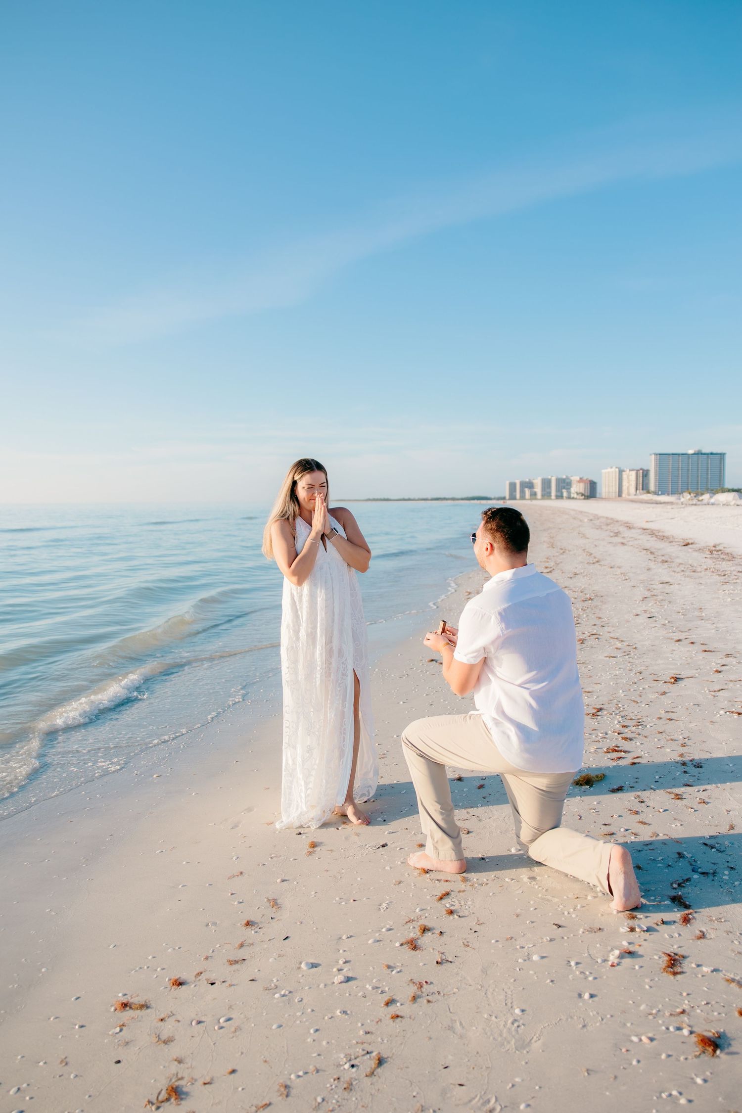 Romantic beach proposal with man on one knee presenting ring to emotional woman by the ocean.