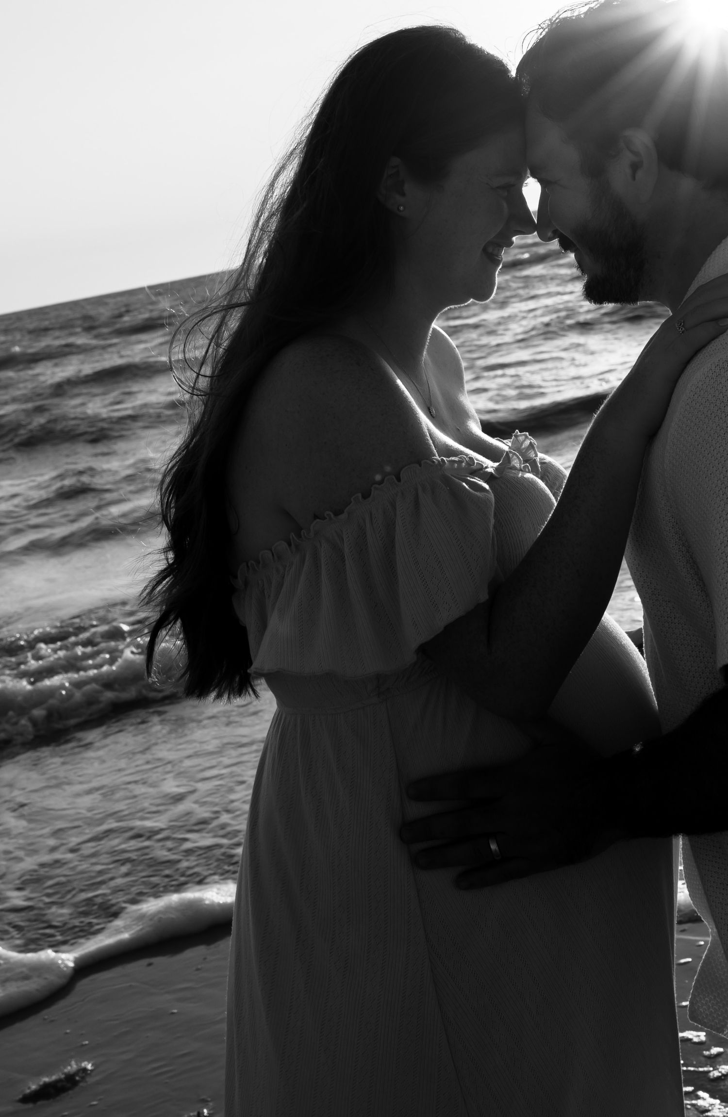Black and white photo of a romantic couple embracing on a beach at sunset, touching foreheads tenderly.