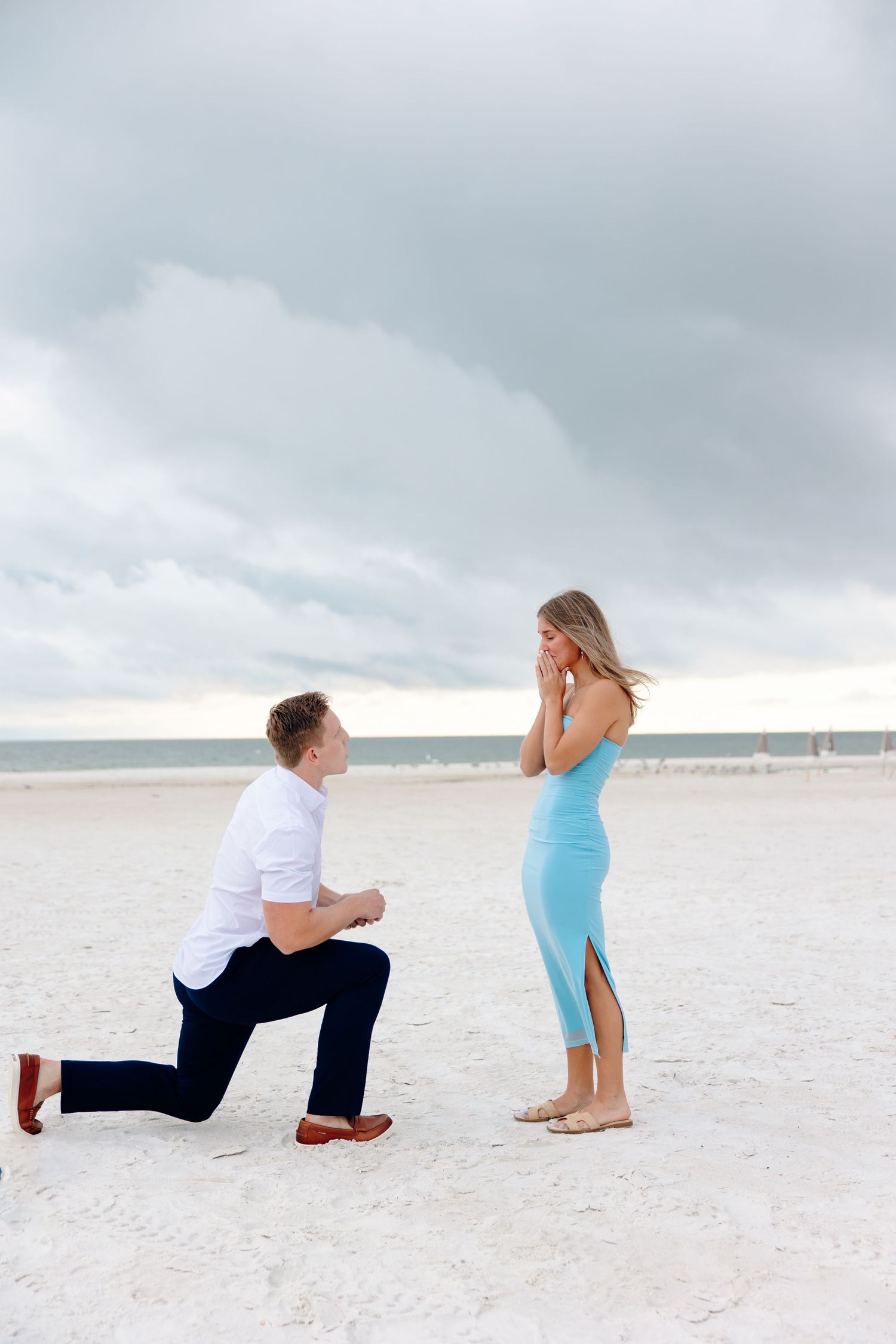 A couple in casual attire performs a surprise beach proposal on white sand under cloudy skies.