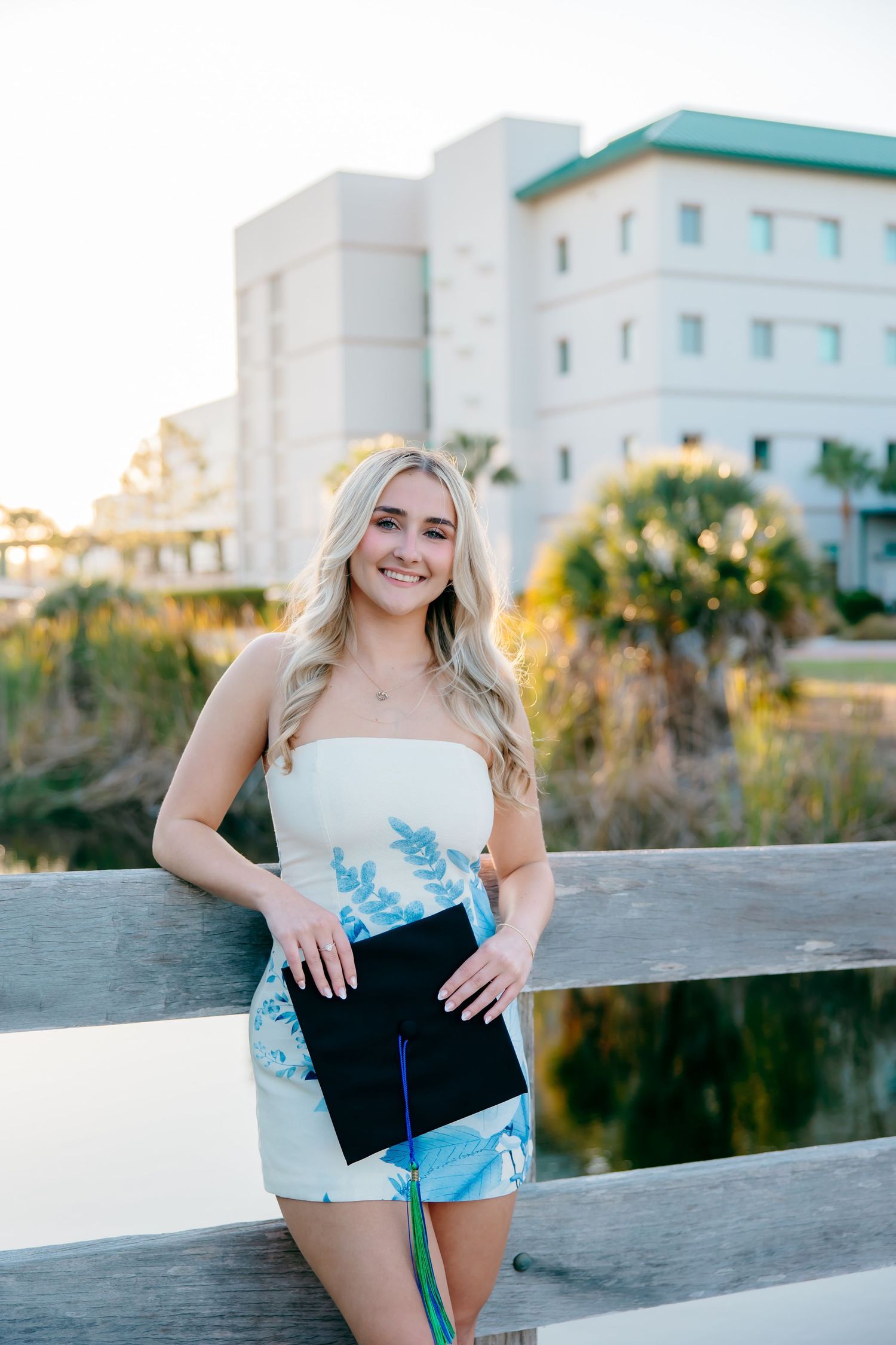 Happy blonde graduate in floral dress holding cap on wooden bridge at sunset.