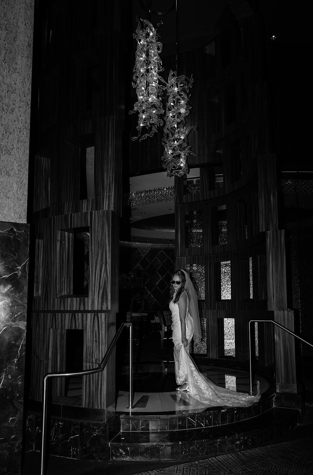 Bride in wedding gown and veil stands on stairs in elegant dark venue with decorative chandelier above.