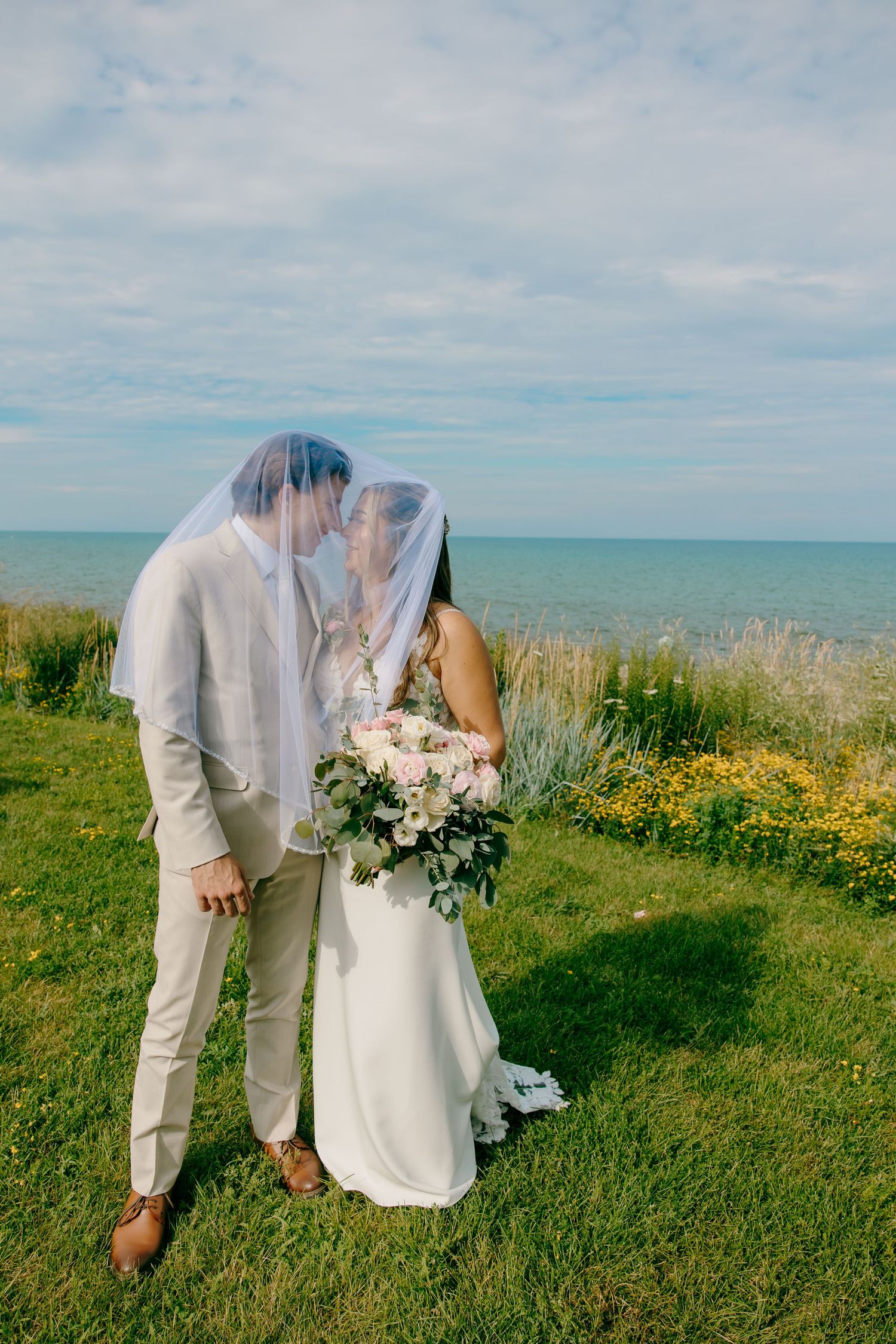 Bride and groom kissing under veil on green cliff overlooking ocean, bride holding white floral bouquet.
