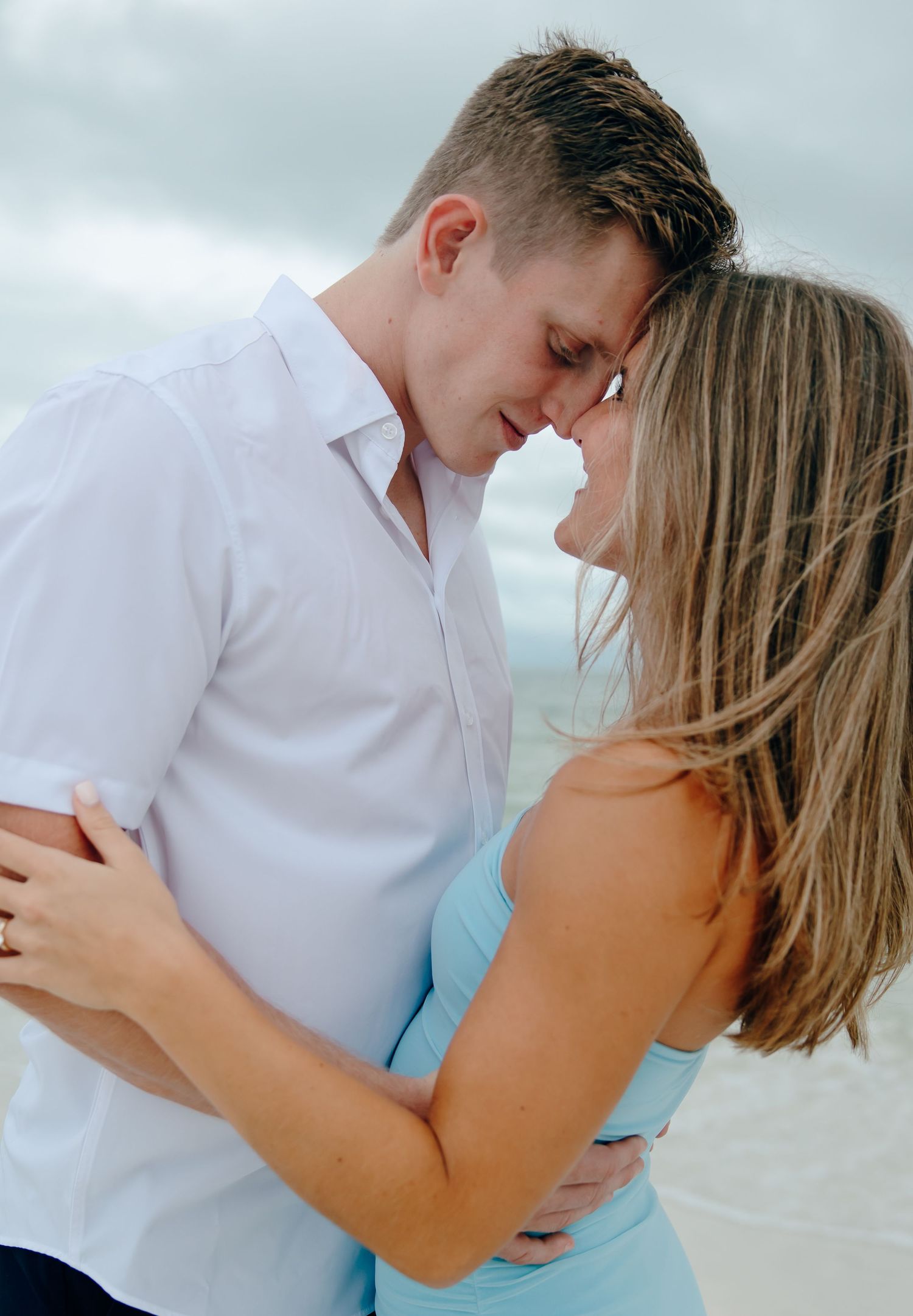A couple in white and blue attire embraces intimately on a beach during a romantic photoshoot sequence.