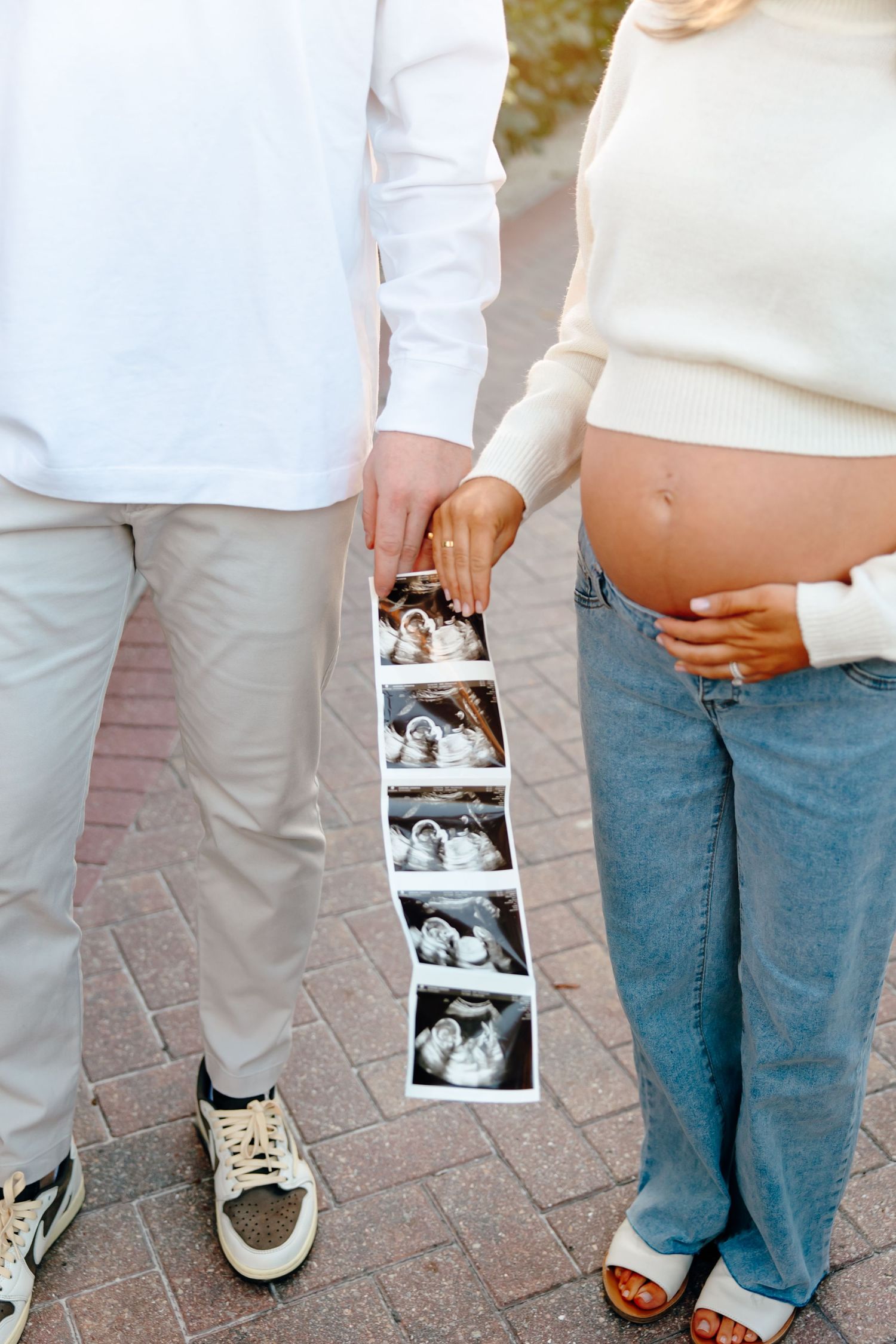 Pregnant woman and partner holding ultrasound photos outdoors, showing baby bump in jeans and white sweater.