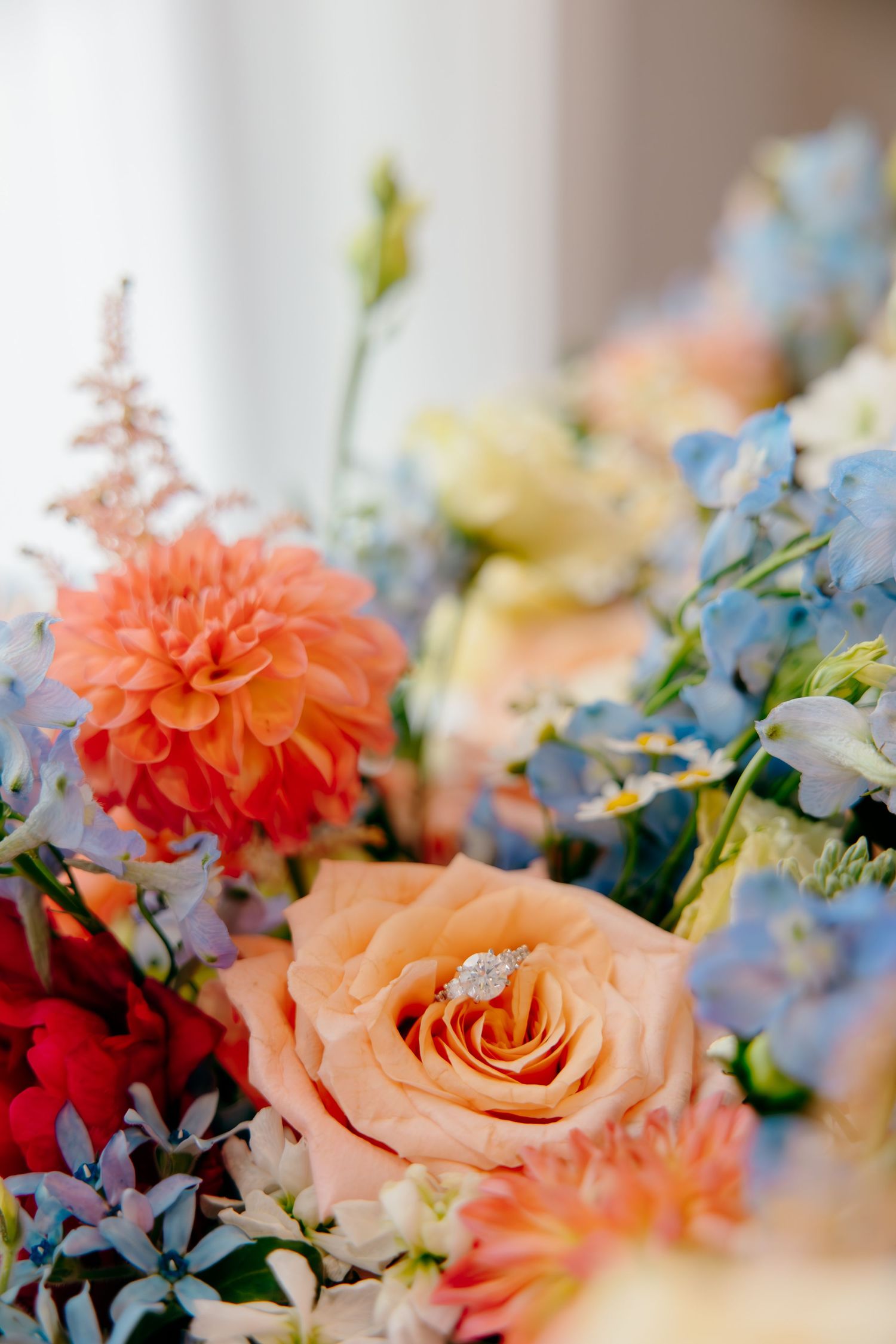 A colorful wedding bouquet featuring peach roses, orange dahlias, and blue flowers with a diamond engagement ring nestled inside.