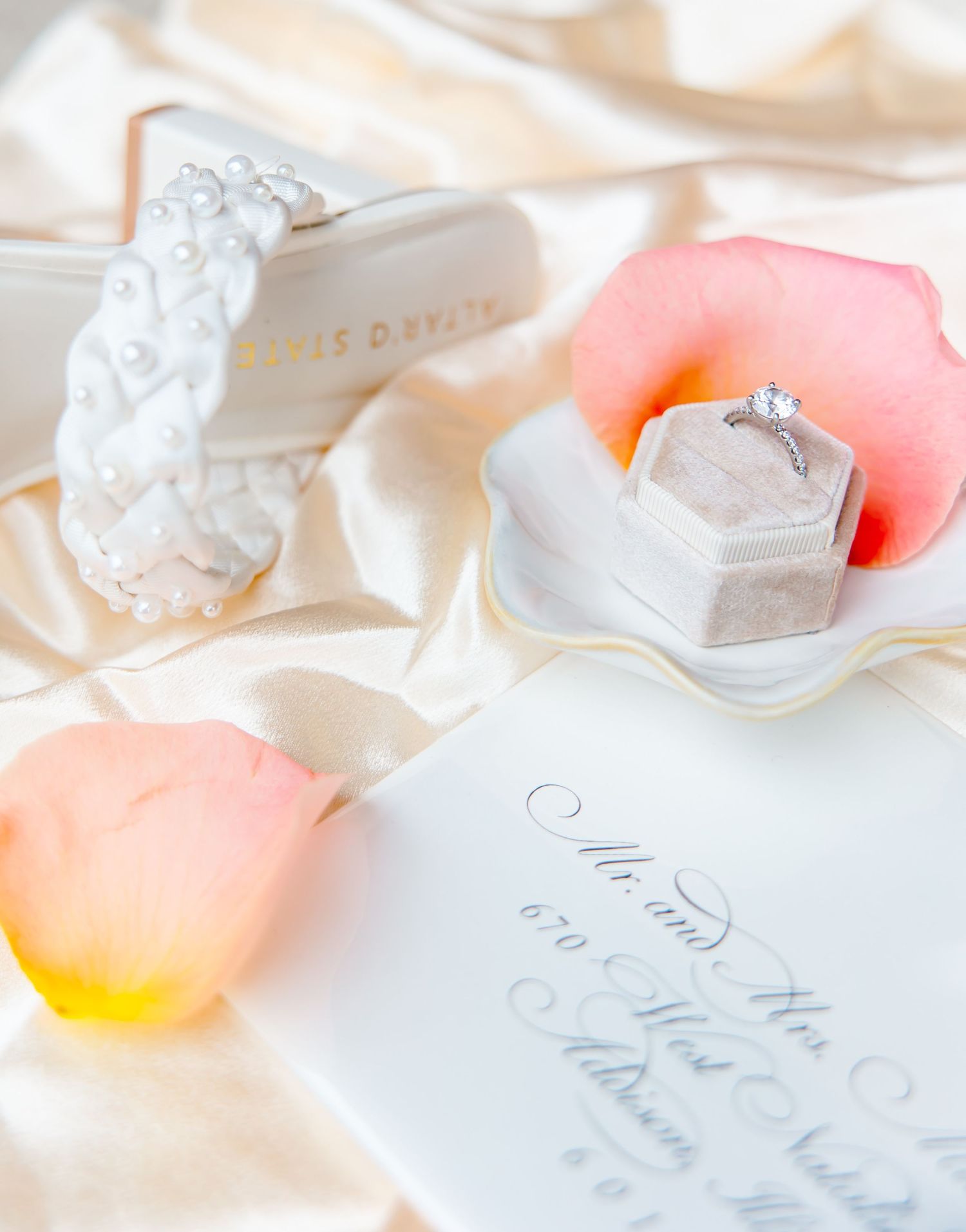 Elegant wedding flat lay with diamond ring in velvet box, pearl bracelet, rose petals, and calligraphy invitation on silk.