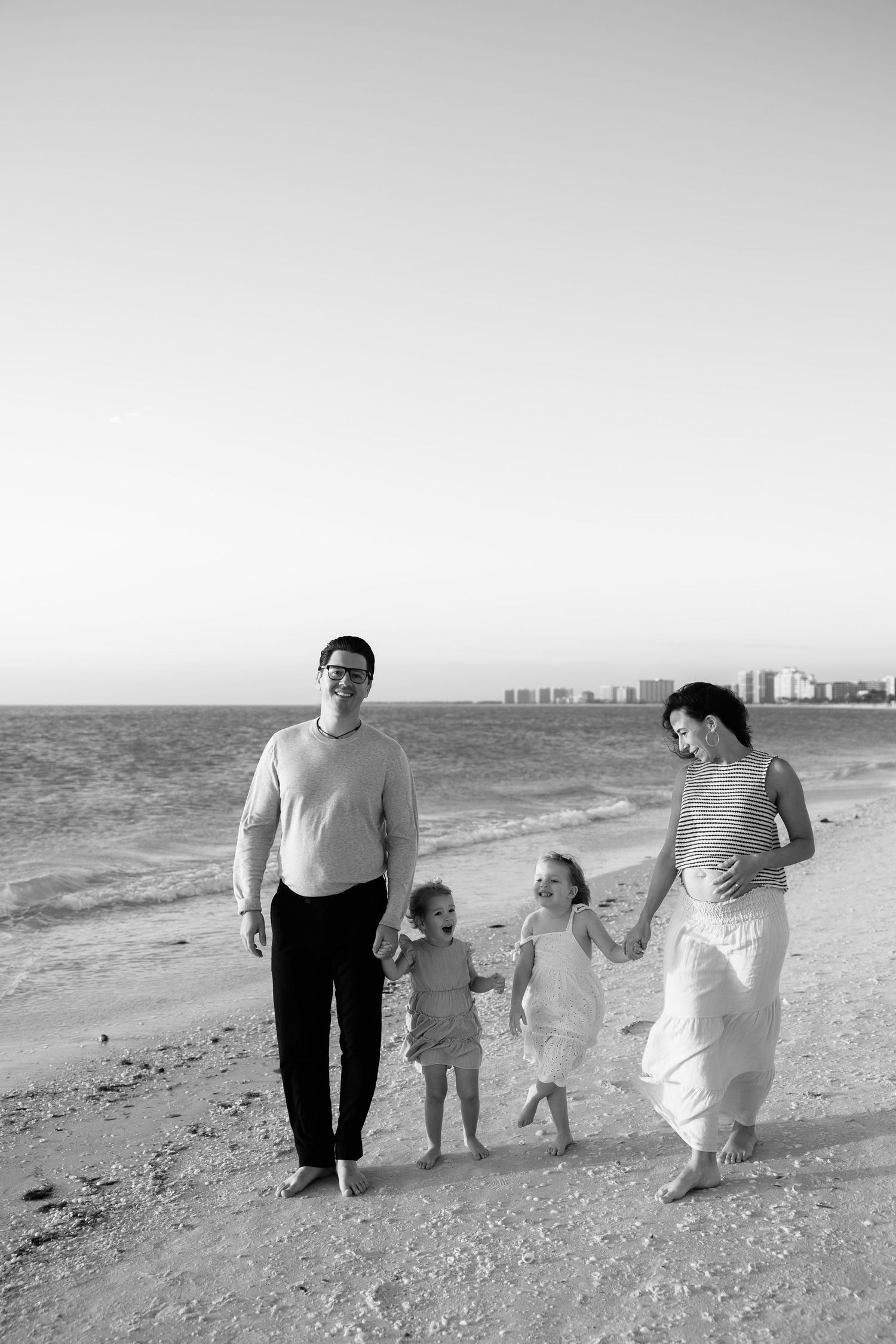 Black and white photo of a family of four walking on a beach, two parents holding hands with two young children.