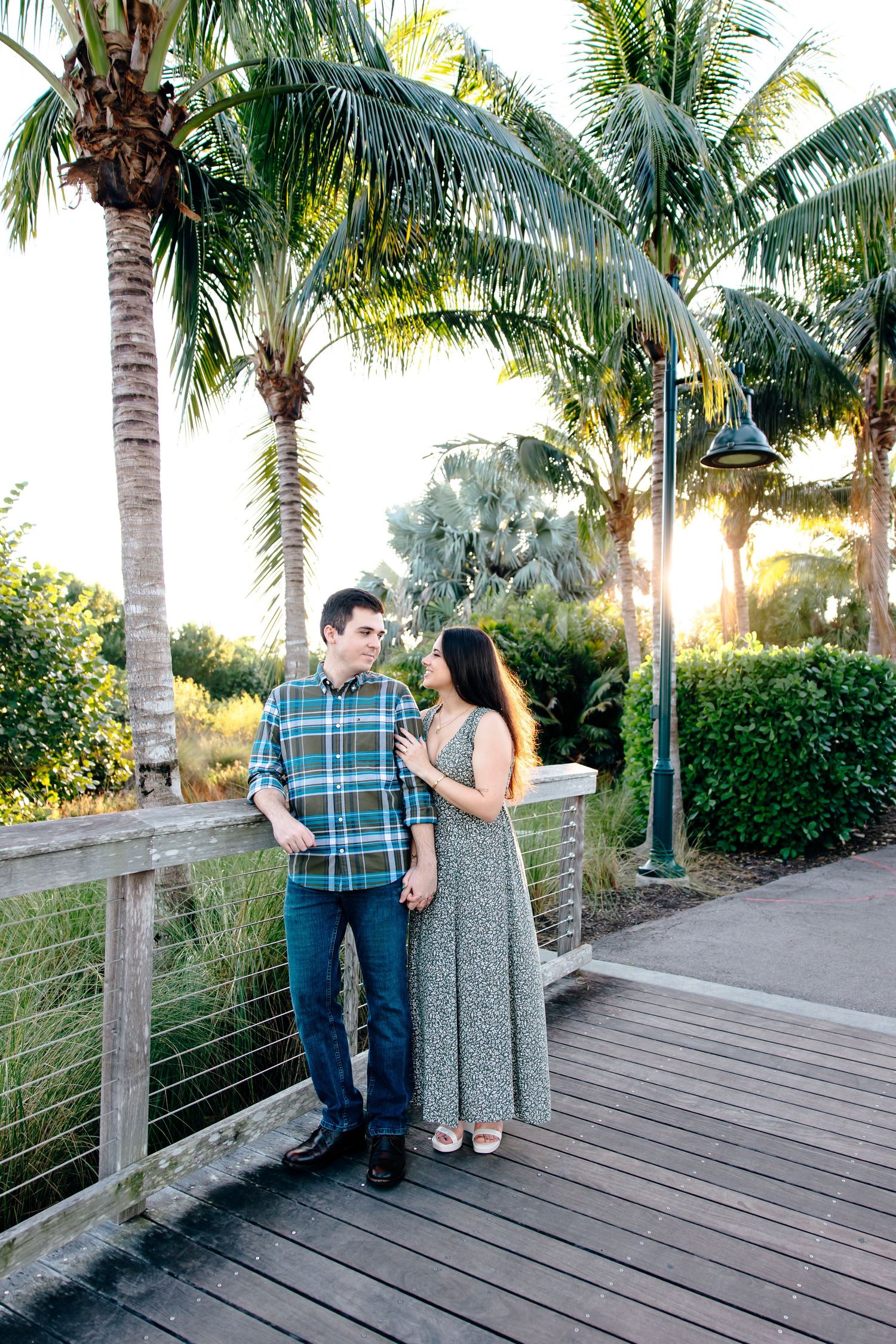 Couple standing on a wooden boardwalk surrounded by palm trees, sharing a romantic moment in a tropical setting.