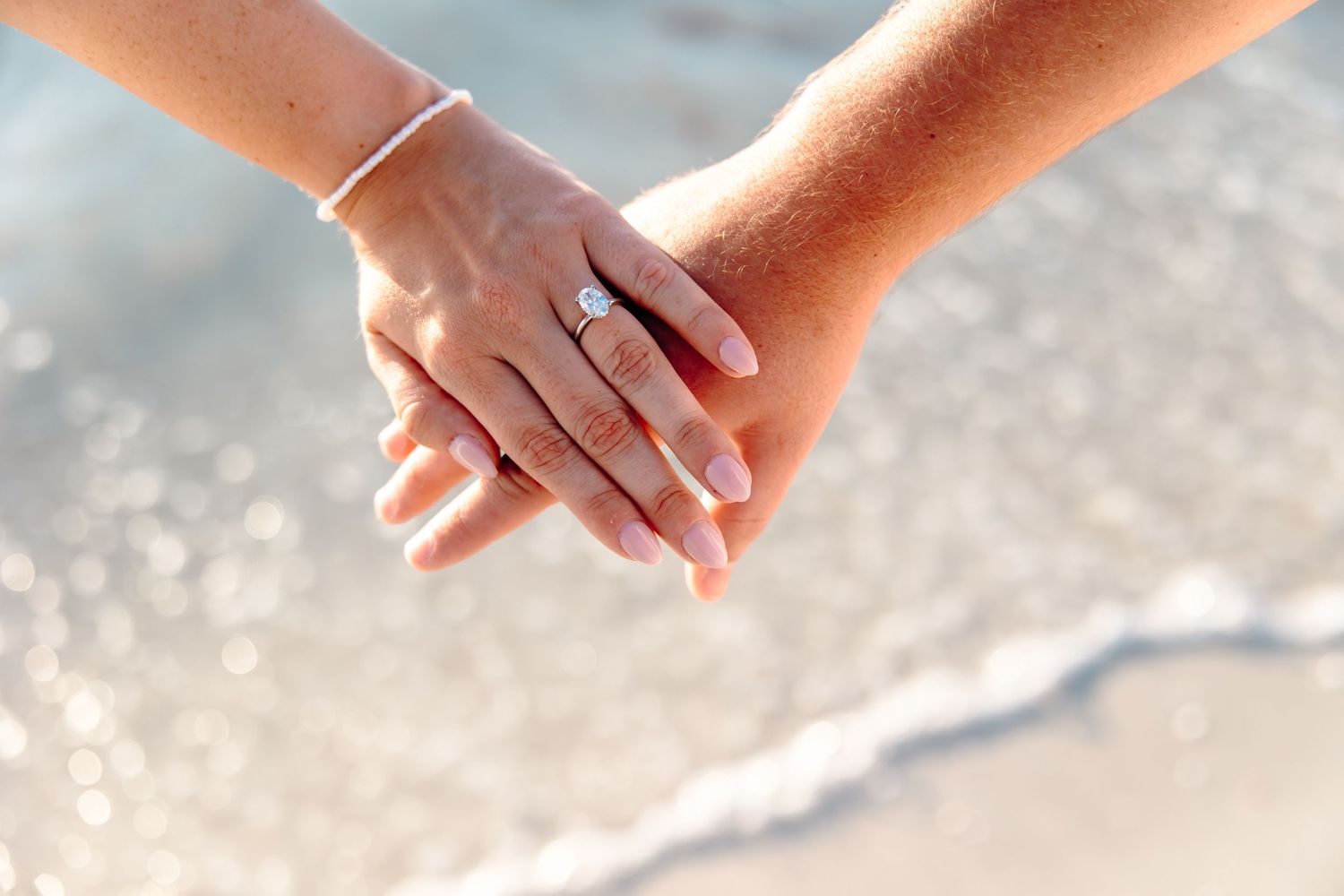 Two hands holding at the beach, woman wearing a diamond engagement ring, ocean waves in the background.