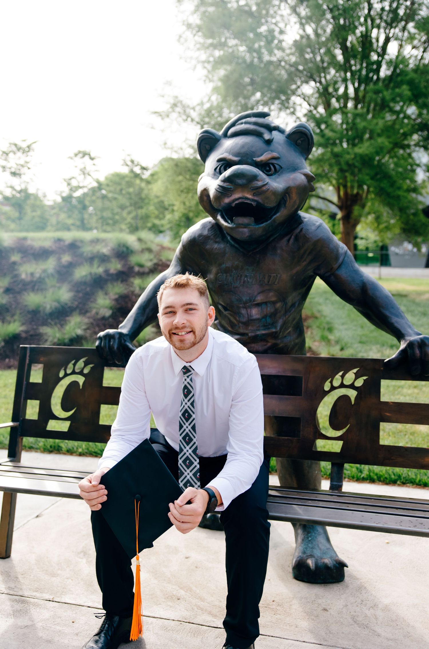 Young man in dress shirt and tie sits on a bench in front of a University of Cincinnati Bearcat statue.