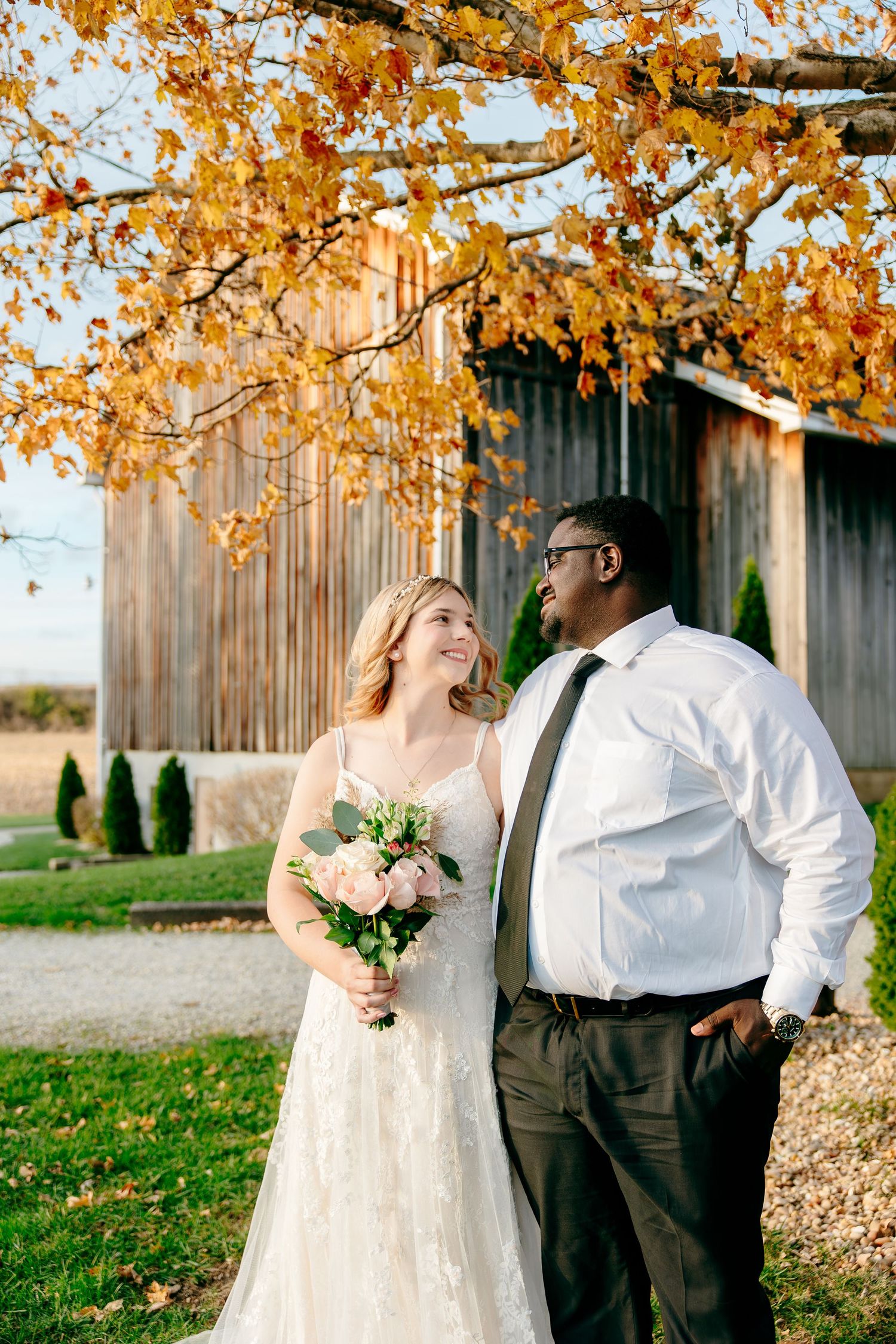 Bride and groom share a joyful moment under autumn foliage near a rustic barn on their wedding day.