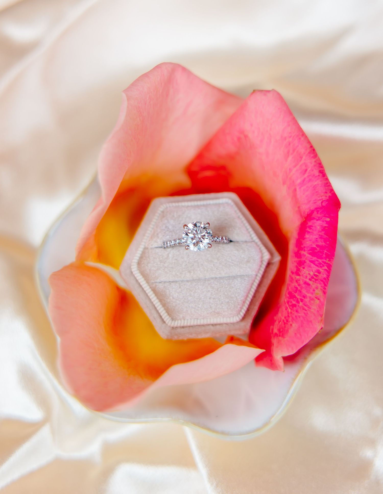 Diamond solitaire engagement ring in hexagonal velvet box nestled inside a pink rose on satin fabric.