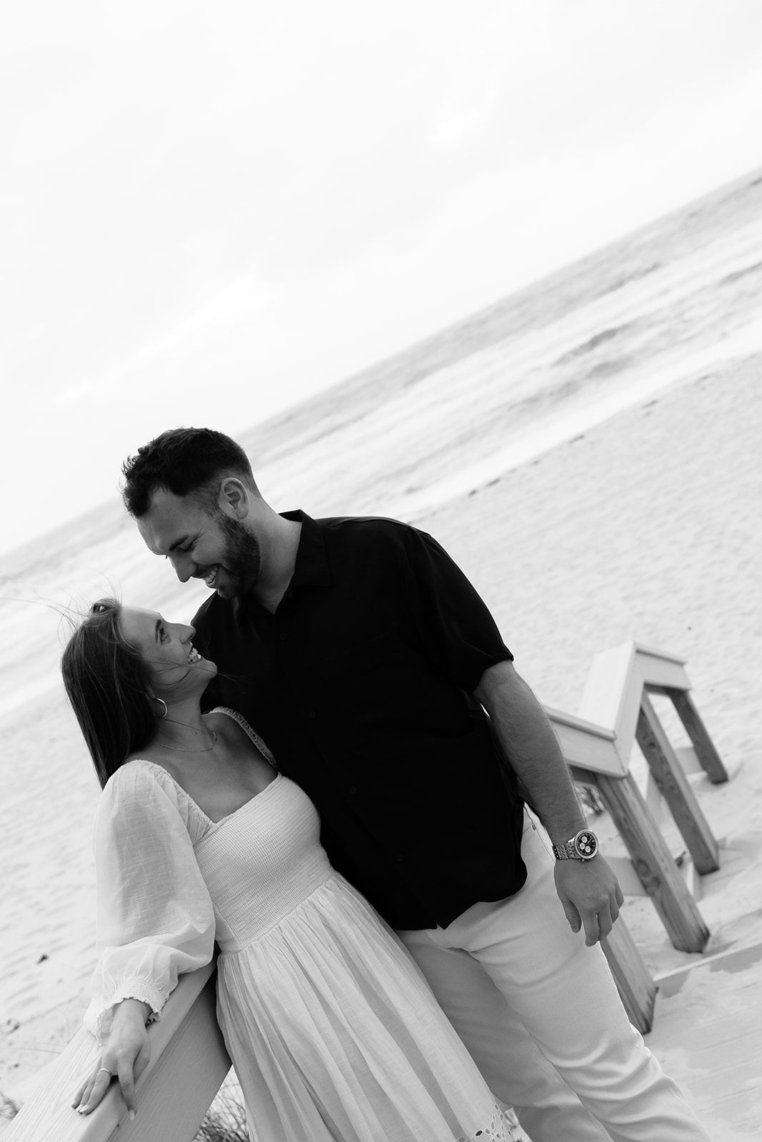 Black and white beach photo of a couple in romantic embrace, woman in white dress, man in black shirt and white pants.