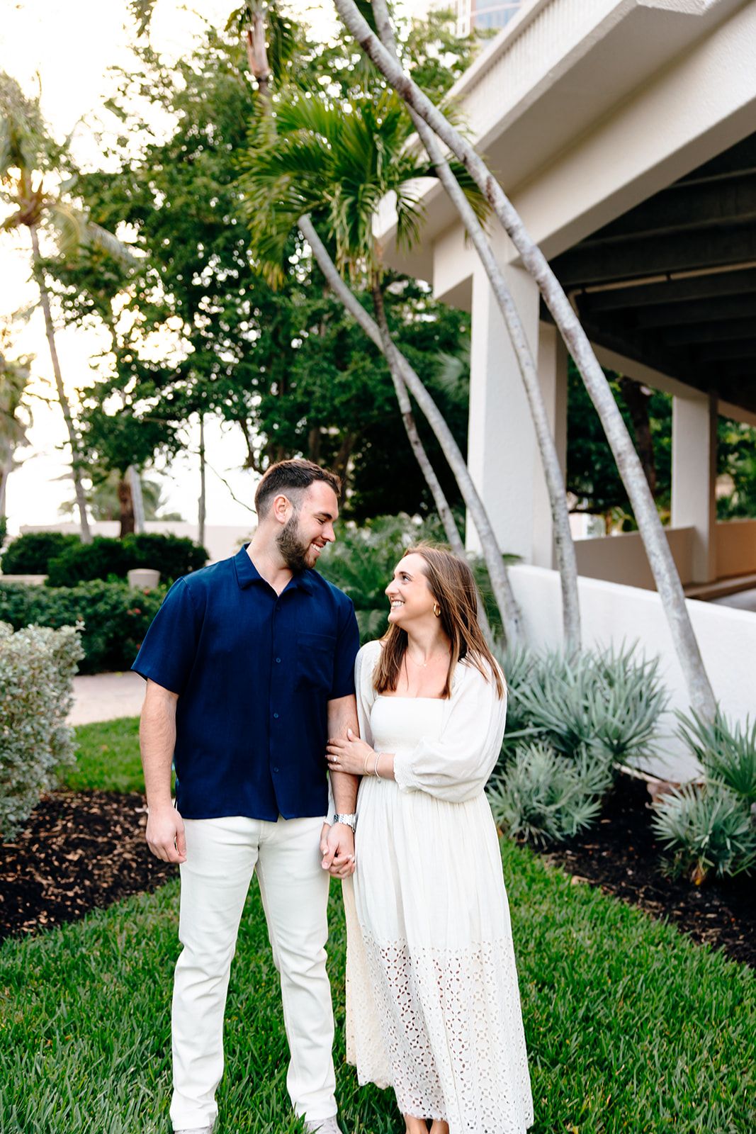 Couple standing together on lawn holding hands, man in navy blue shirt and woman in elegant white dress.