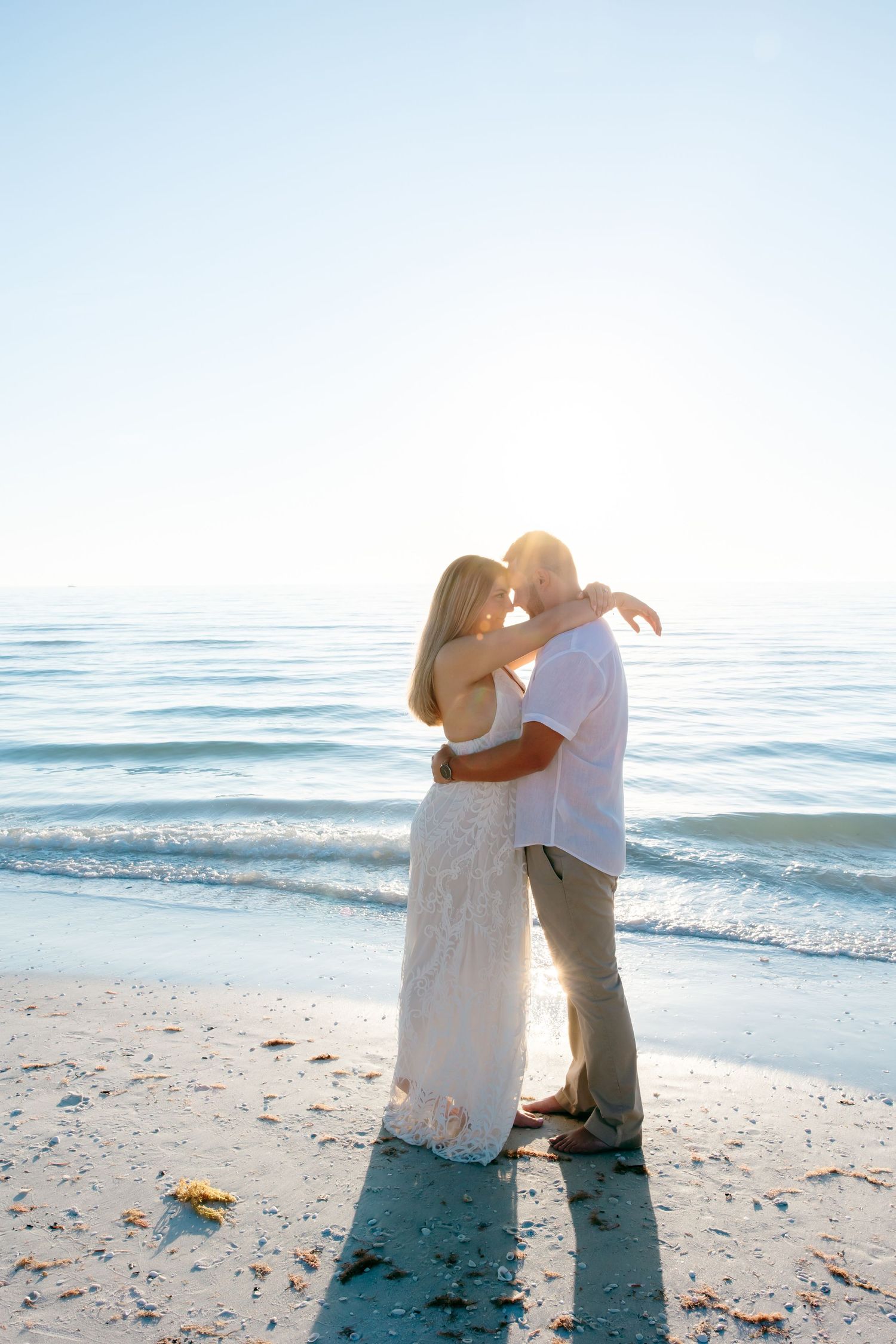 A couple embraces on a sunny beach with ocean waves in the background while sharing a romantic moment.