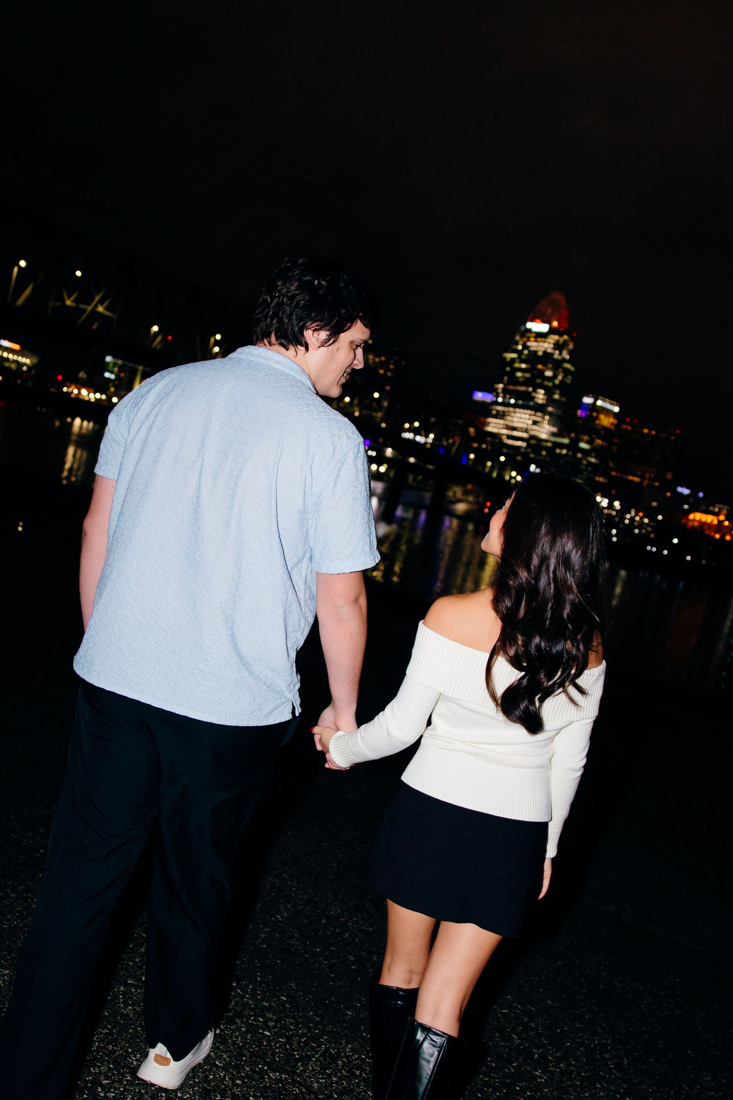 Couple holding hands walking at night with illuminated city skyline reflecting on water in background.