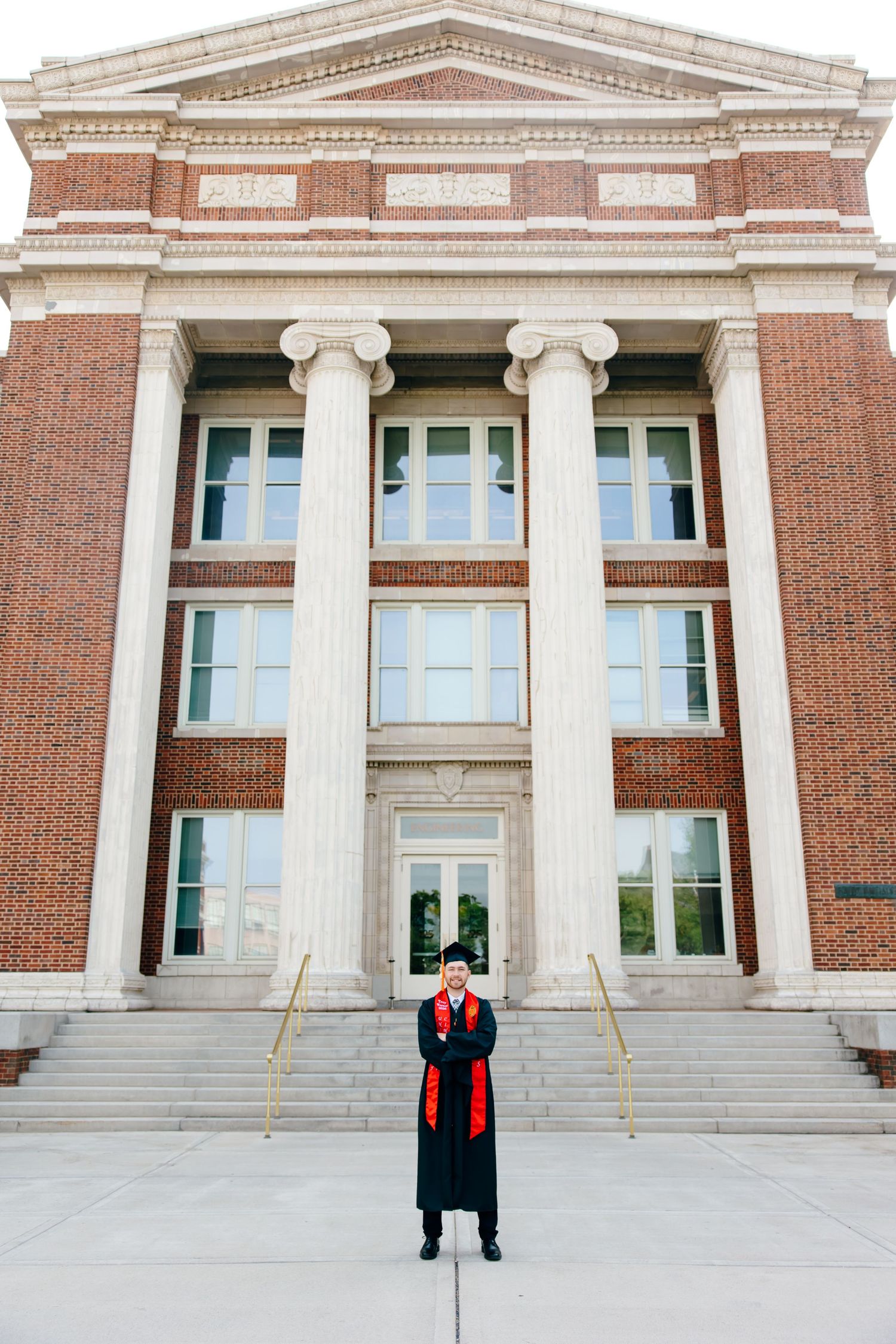 Graduate in black cap and gown with red sash stands before a classic brick university building with white columns.
