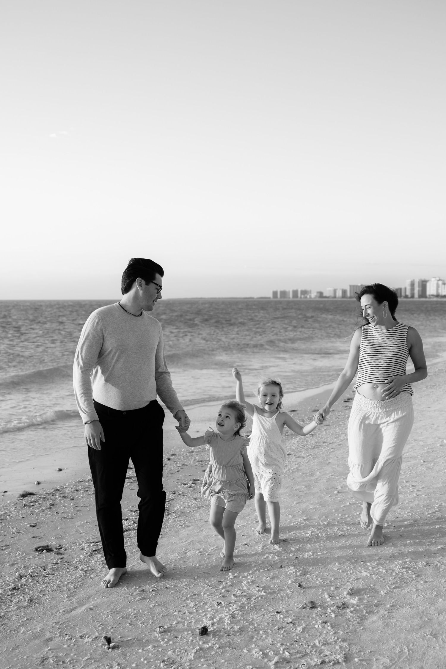 Black and white photo of a happy family of four walking on the beach, parents swinging two young children by their hands.