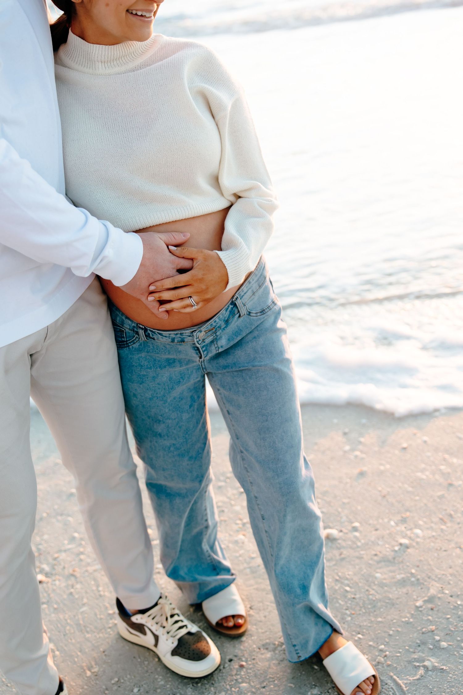 Couple standing on beach, woman in white sweater and jeans with partner's hands on her pregnant belly near ocean waves.
