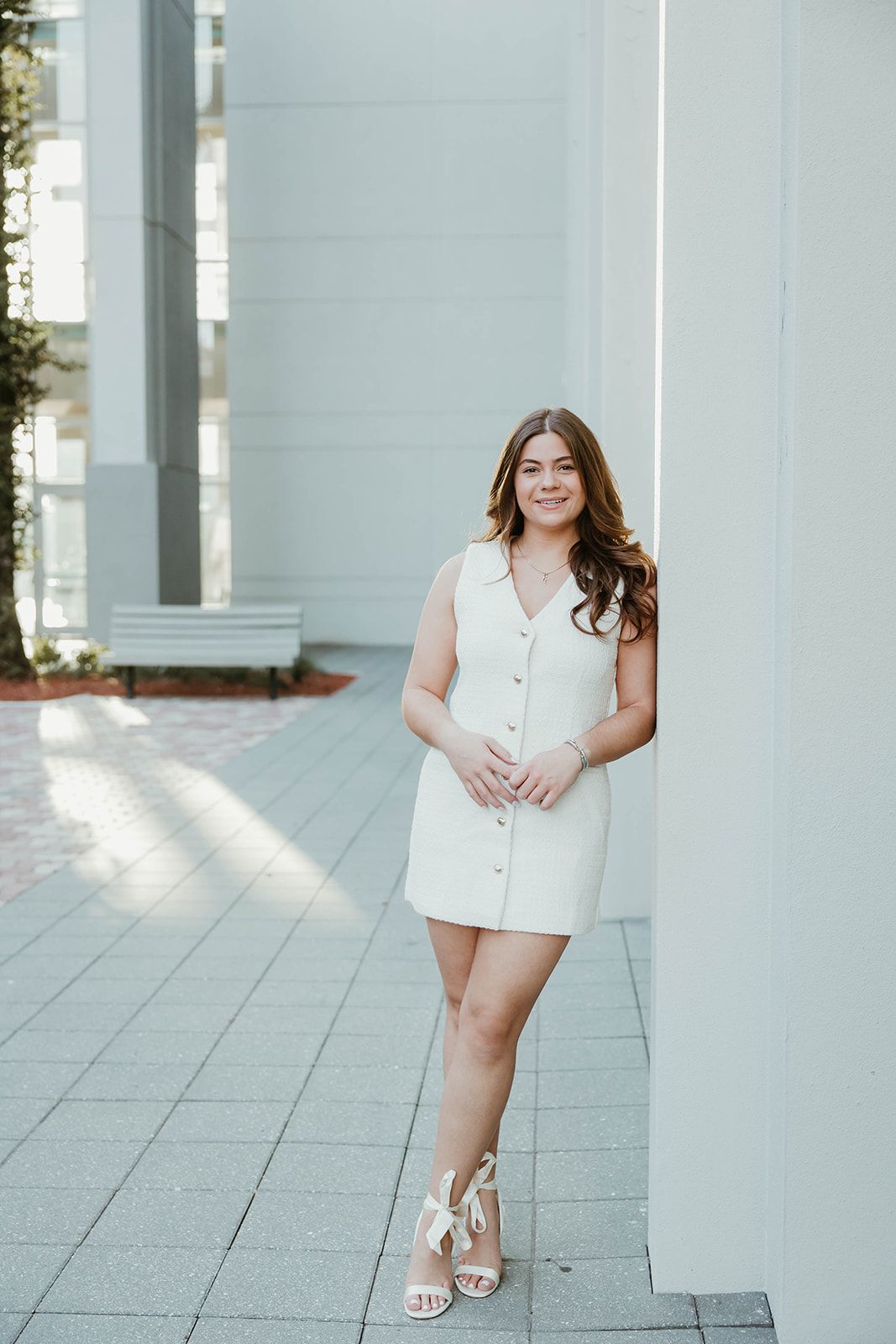A person in a white sleeveless dress poses on a concrete walkway near white columns in natural lighting.