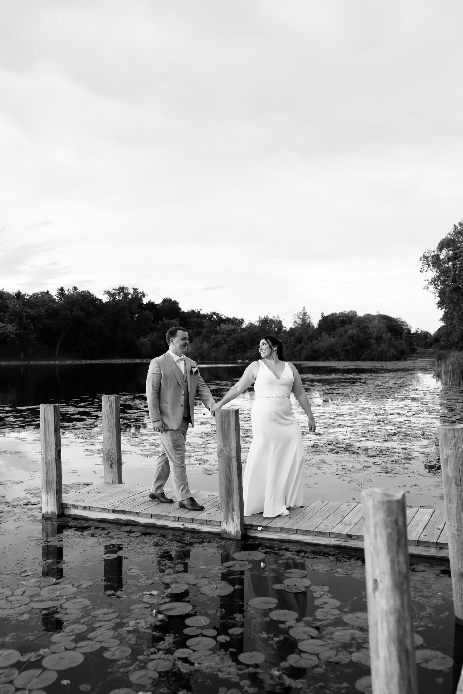 Bride and groom hold hands on a wooden dock by a lily pad lake in a romantic black and white wedding photo.
