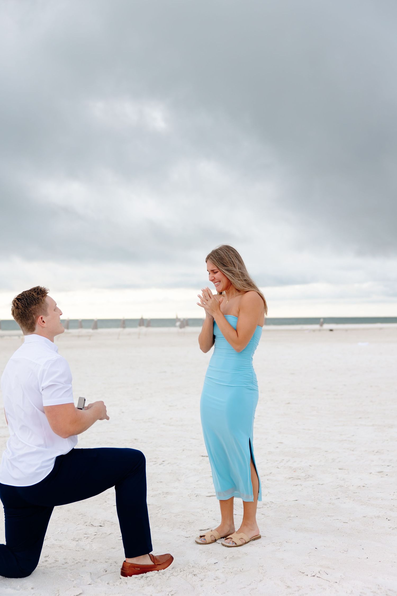 Man proposes on one knee to woman in blue dress on white sand beach under cloudy sky.