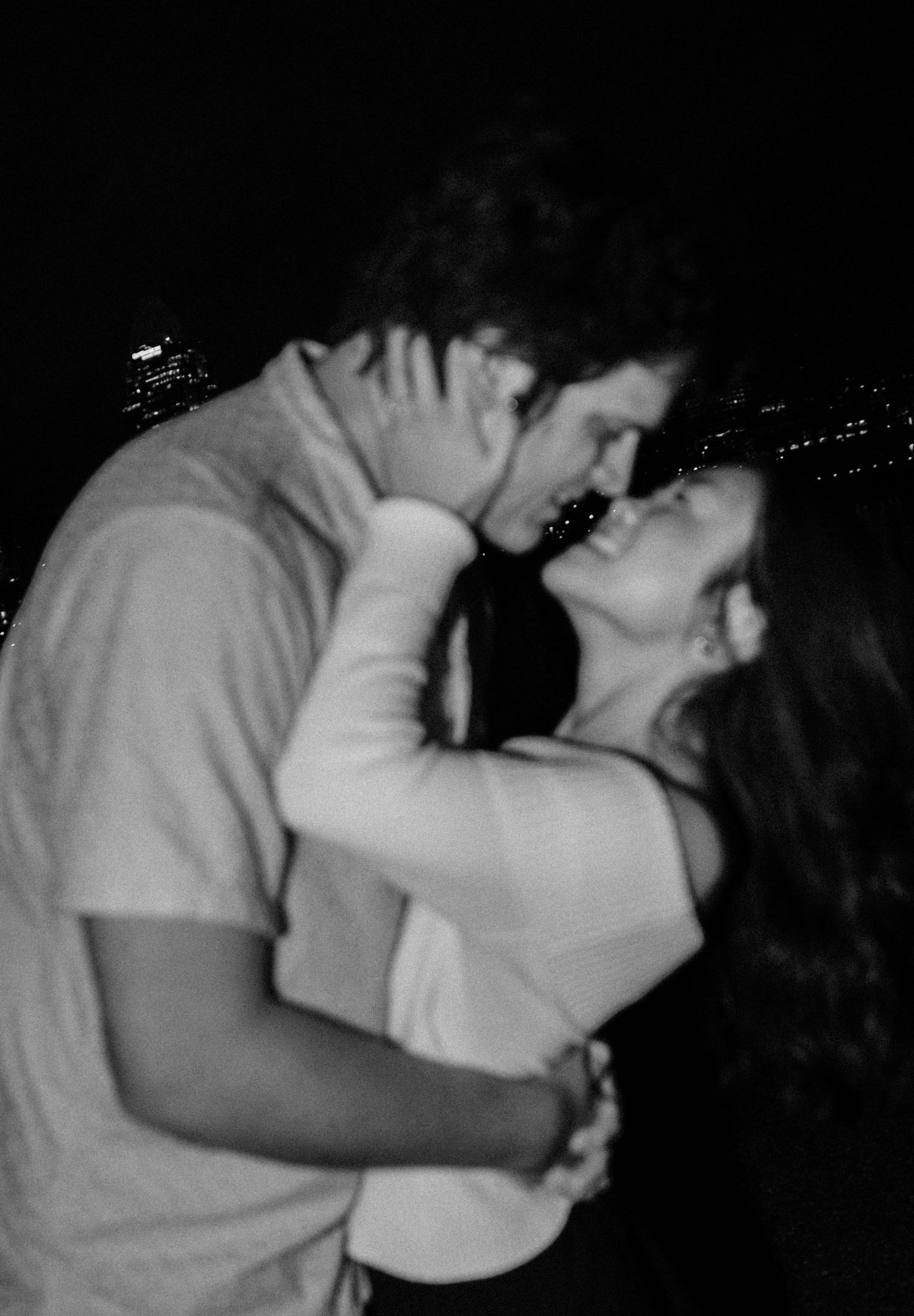Black and white photo of a romantic couple embracing and kissing at night with city lights in the background.