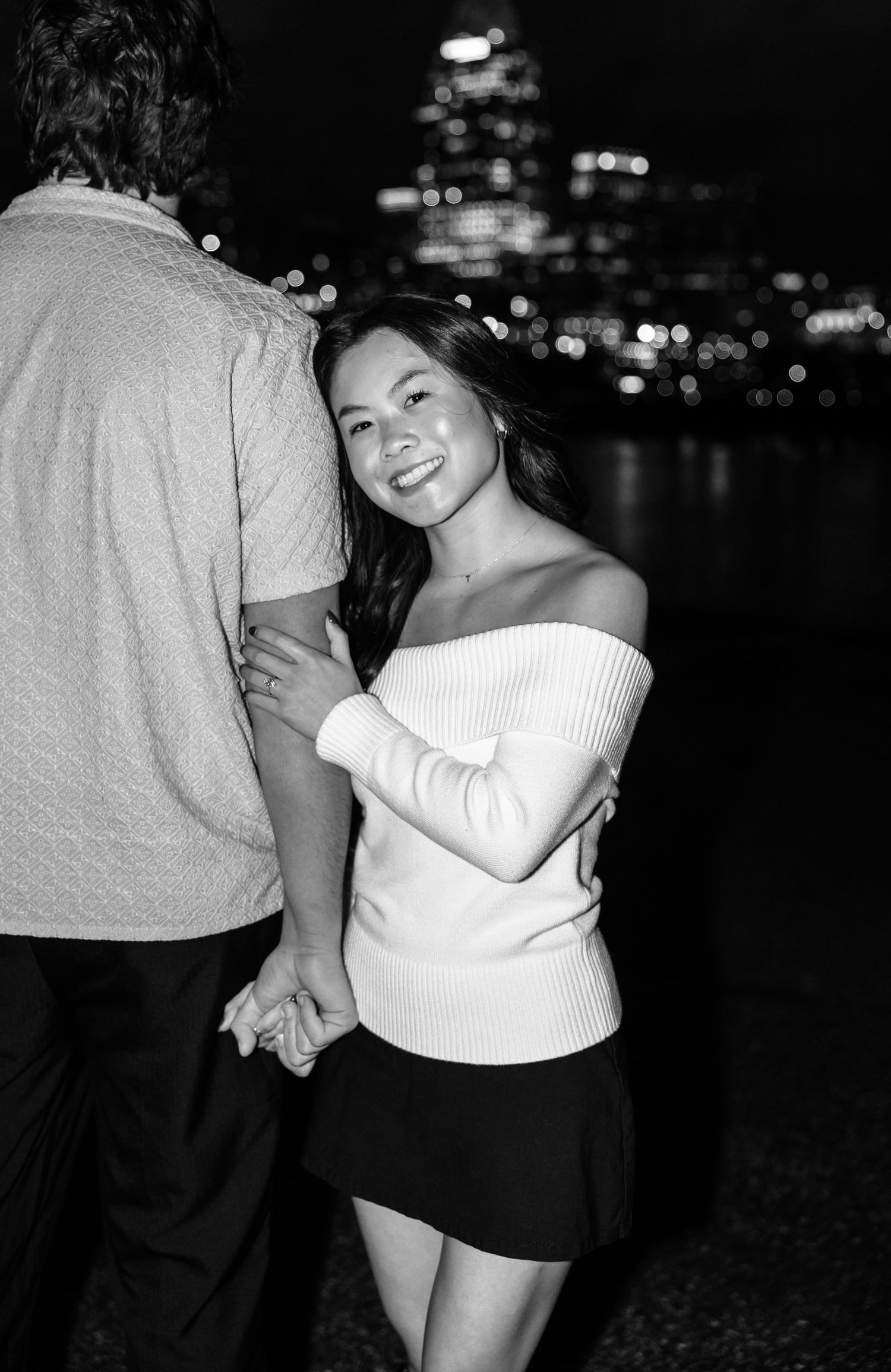 Black and white photo of a smiling woman in off-shoulder top holding hands with a man, city lights glowing behind them.