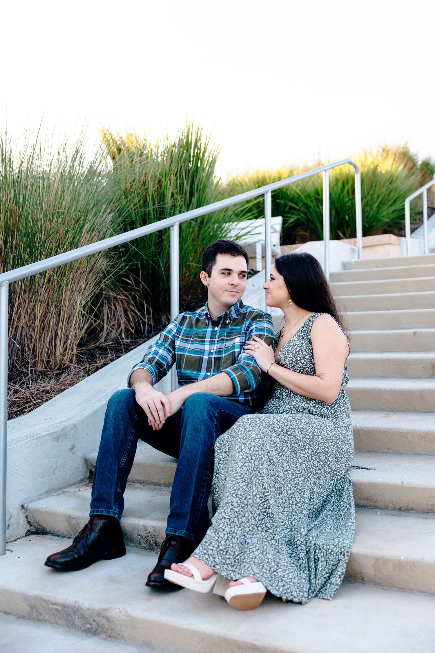 Couple sitting on outdoor steps, woman in floral dress leaning toward man in plaid shirt and jeans, surrounded by tall grass.