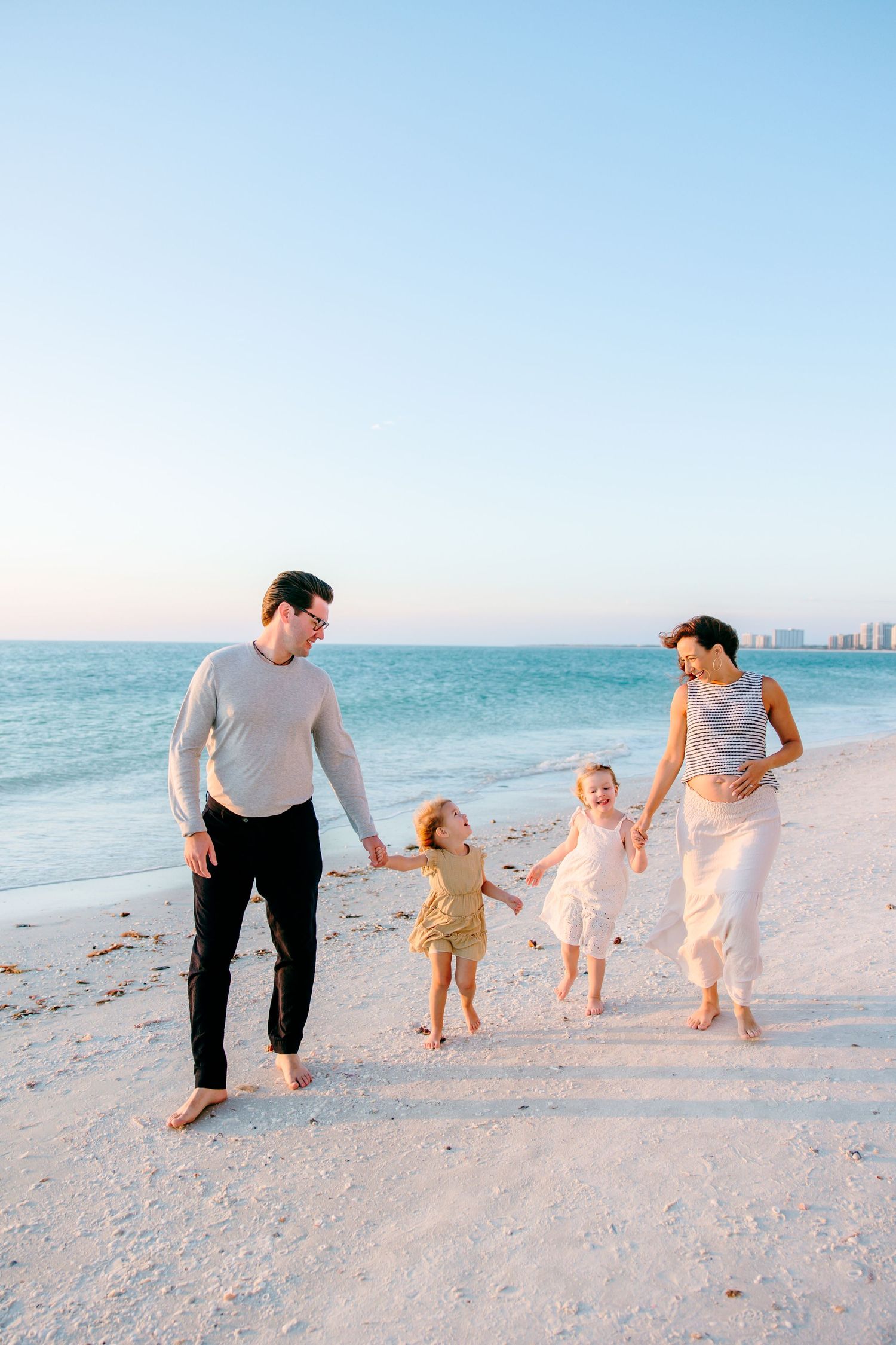 Family of four walking hand-in-hand on a sandy beach at sunset, with calm ocean waves and city skyline in background.