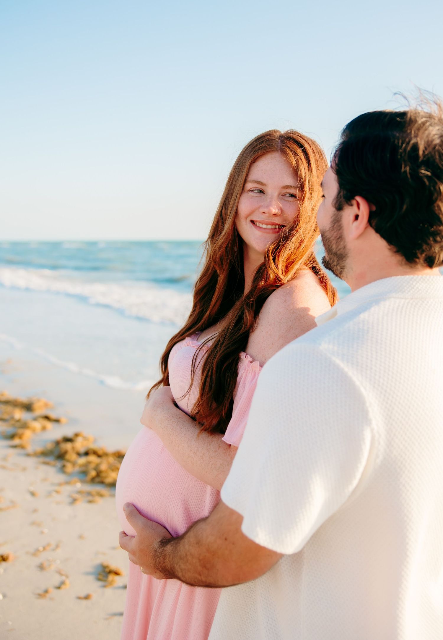 Pregnant woman in pink dress smiles at partner on sunny beach, waves and blue sky in background.