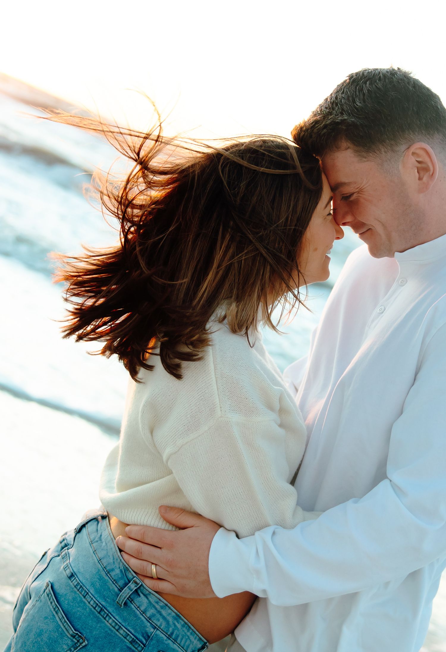 Romantic couple embracing on a windy beach, foreheads touching, woman's hair blowing in breeze, wearing casual white sweaters.