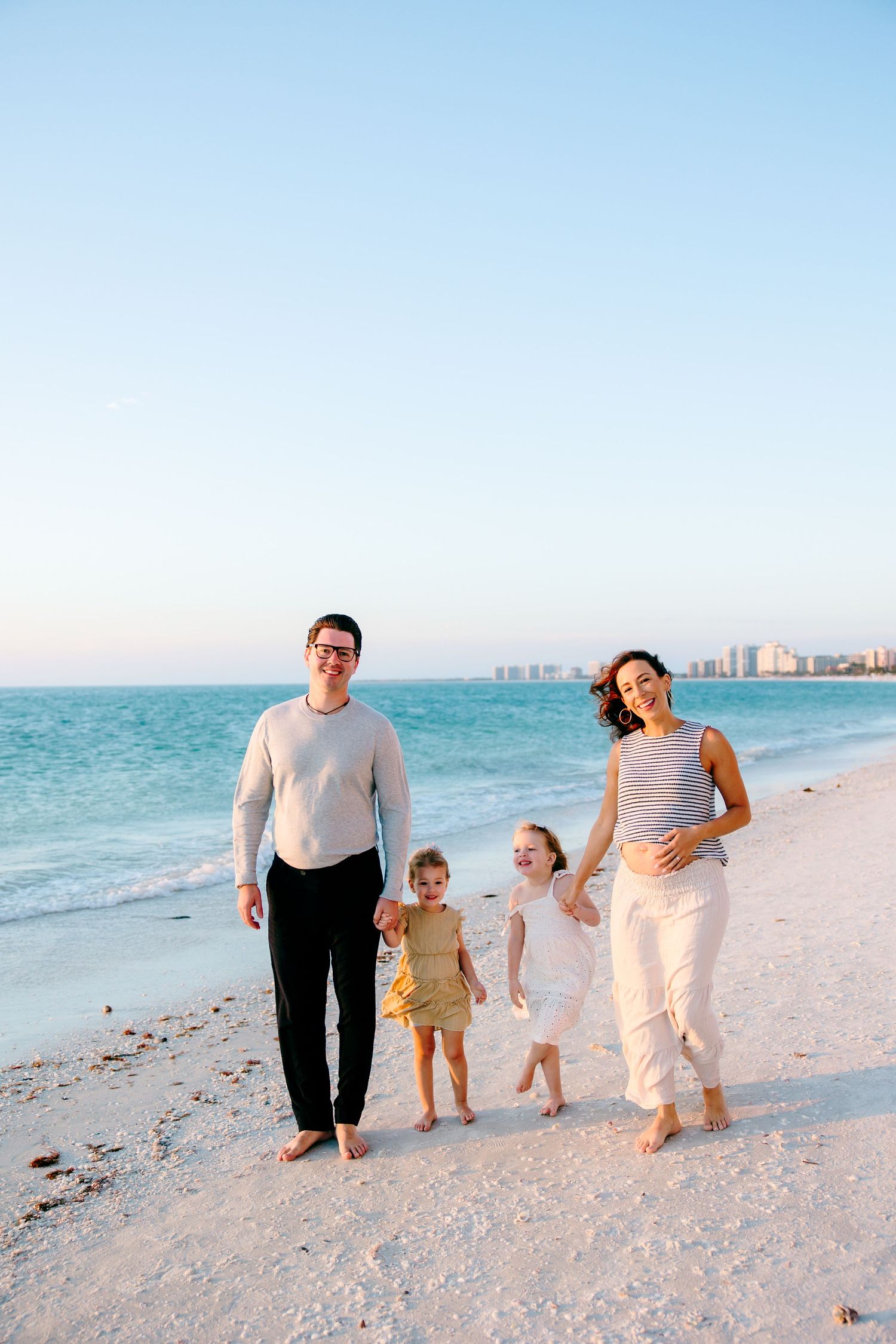Happy family of four walks barefoot on a sunny Florida beach, with turquoise water and city skyline in background.