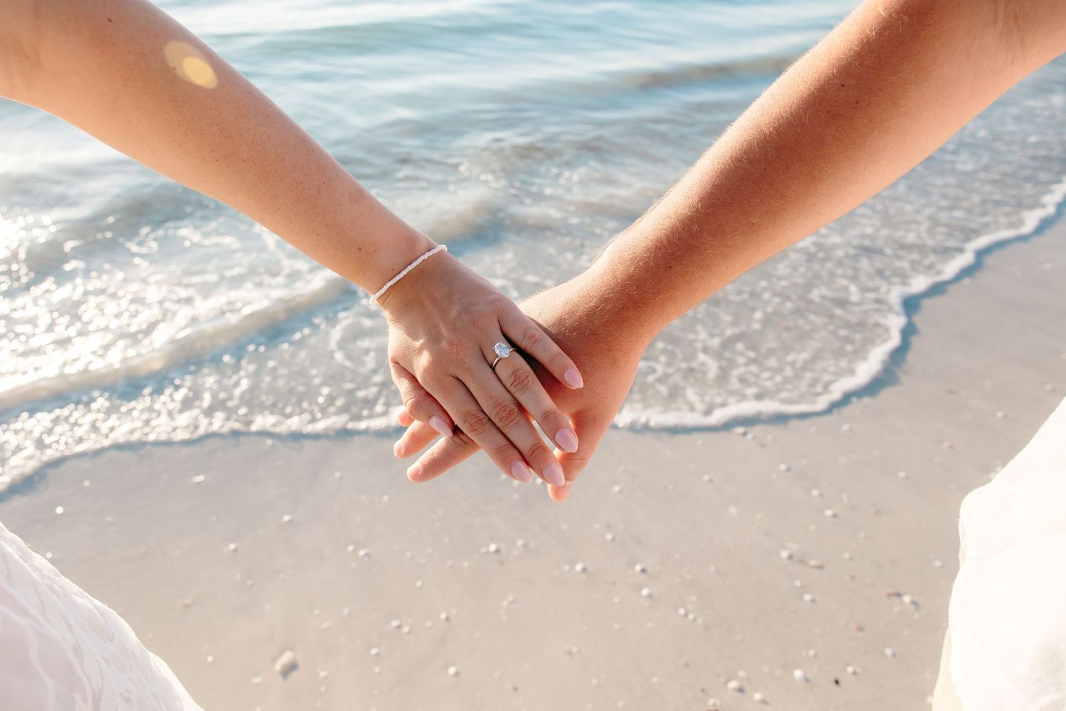 Couple holding hands on a sandy beach with gentle ocean waves, woman wearing an engagement ring.