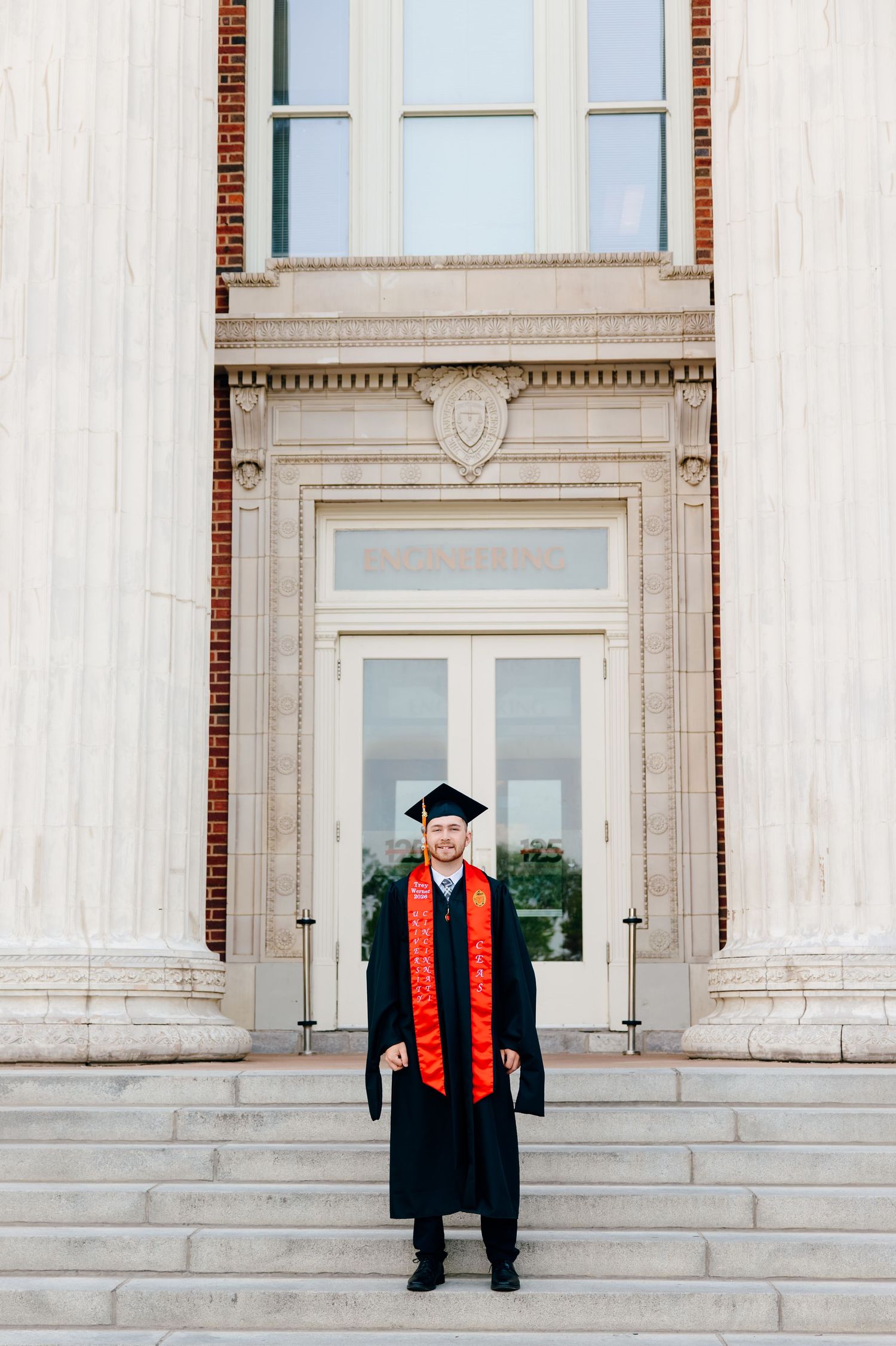 A proud graduate in cap and gown with red sash stands on steps before a grand university building entrance.