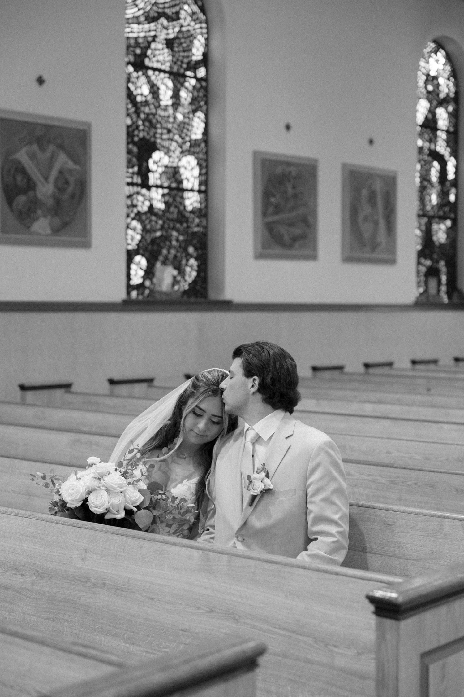 Black and white photo of a bride and groom sharing an intimate moment in a church pew with stained glass windows.