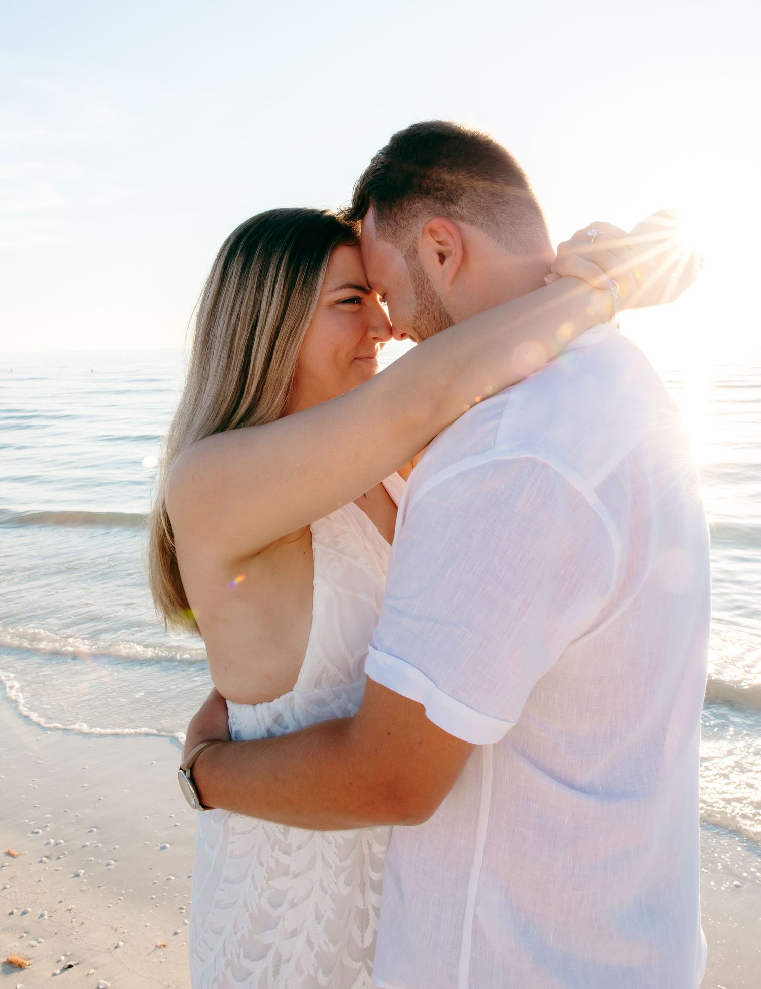 Couple embracing and almost kissing on a sunny beach, wearing white outfits with ocean waves in the background.