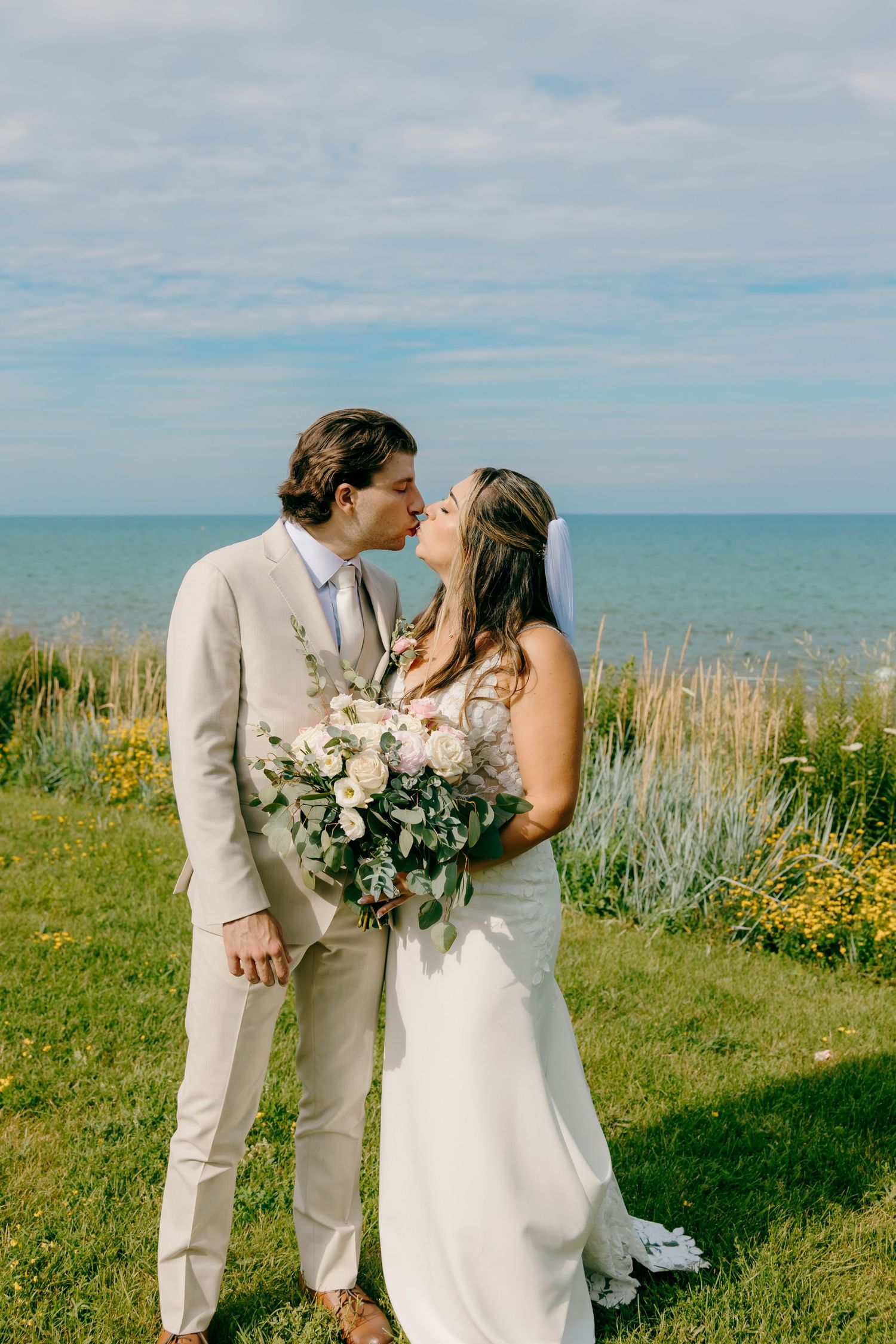 Bride and groom share a kiss outdoors near the ocean, she holds a white bouquet, he wears a tan suit.