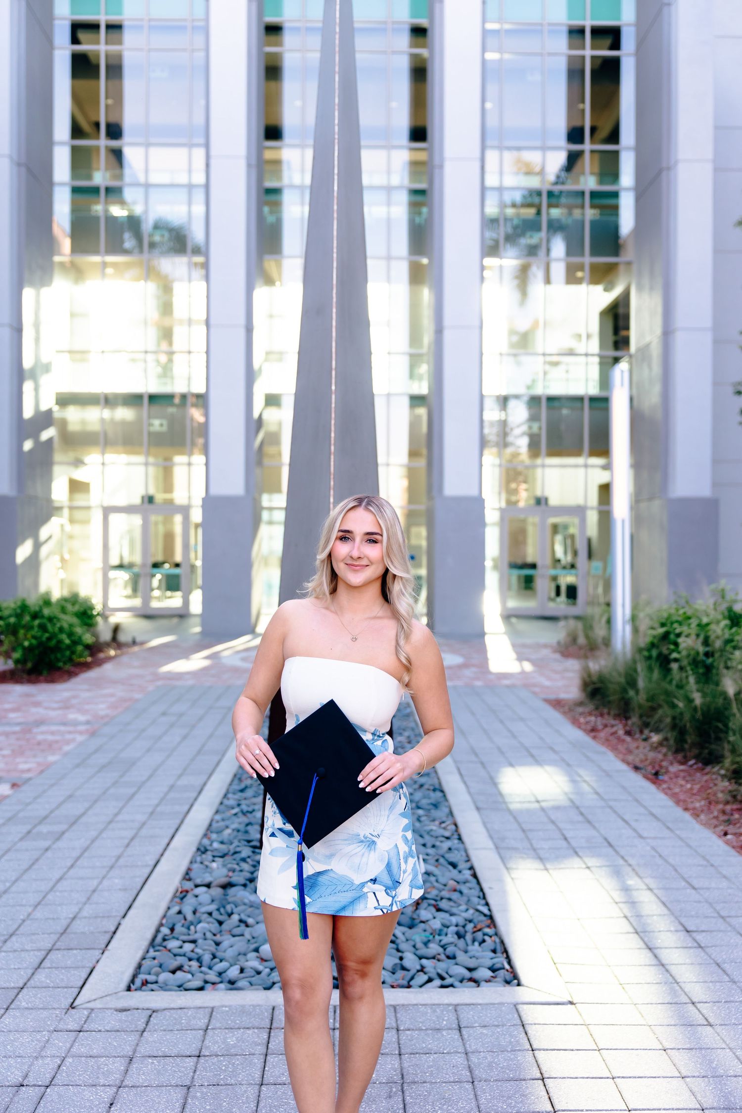 Smiling blonde graduate in floral dress holding cap outside modern university building.
