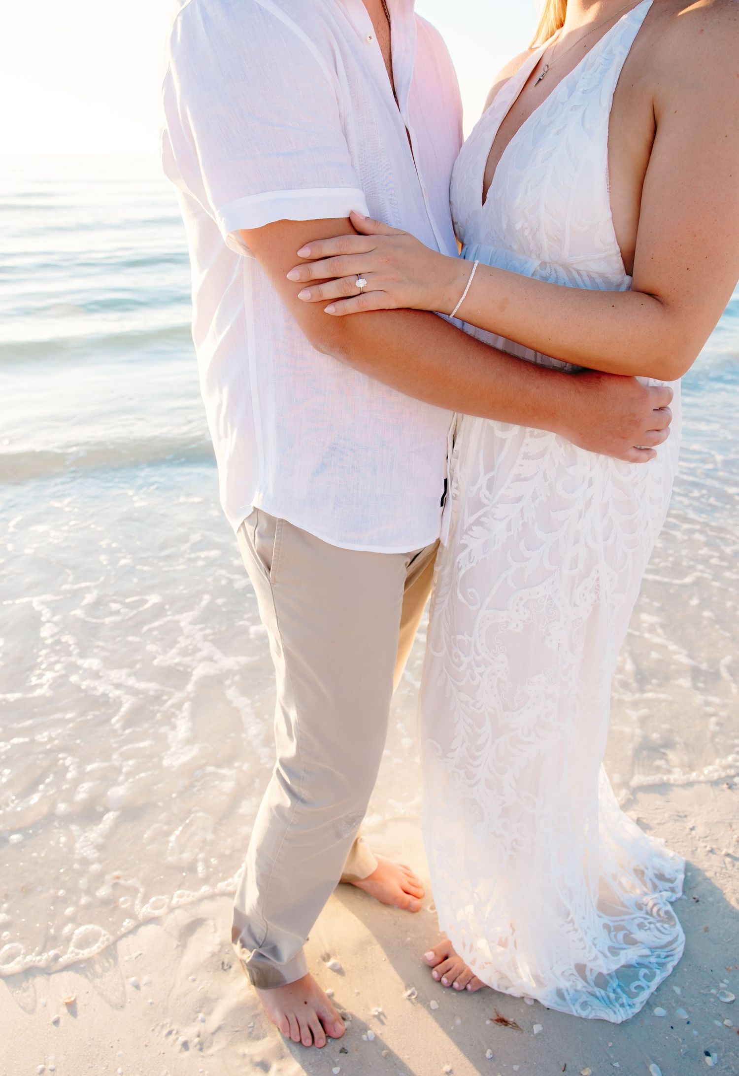 Couple in white beach attire embracing on sandy shore with ocean waves in the background during sunset.