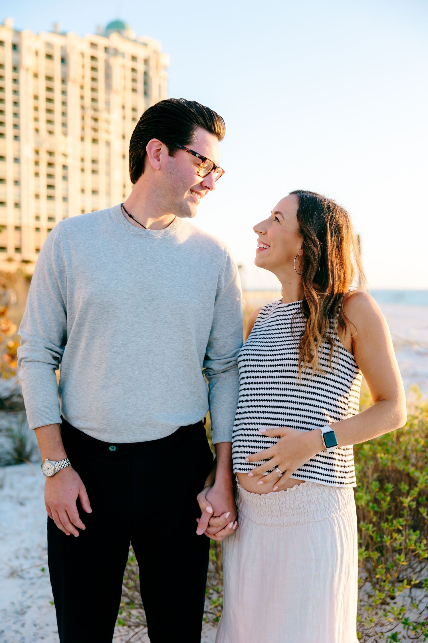 Couple holding hands on beach, woman cradling baby bump in striped dress, man in gray sweater smiling at her.