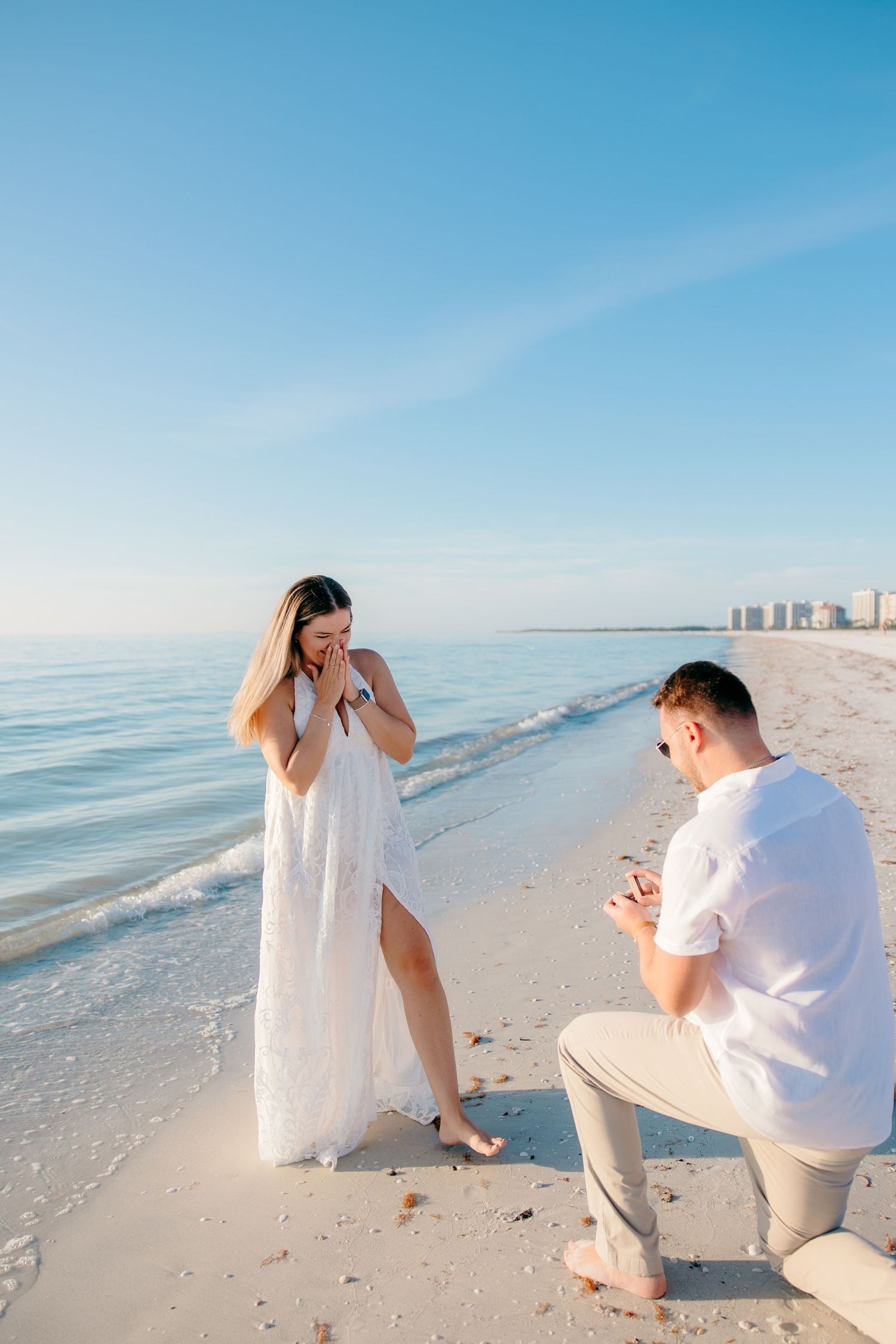 Man proposing to woman on a sunny beach, she reacts with surprise while he kneels with a ring.