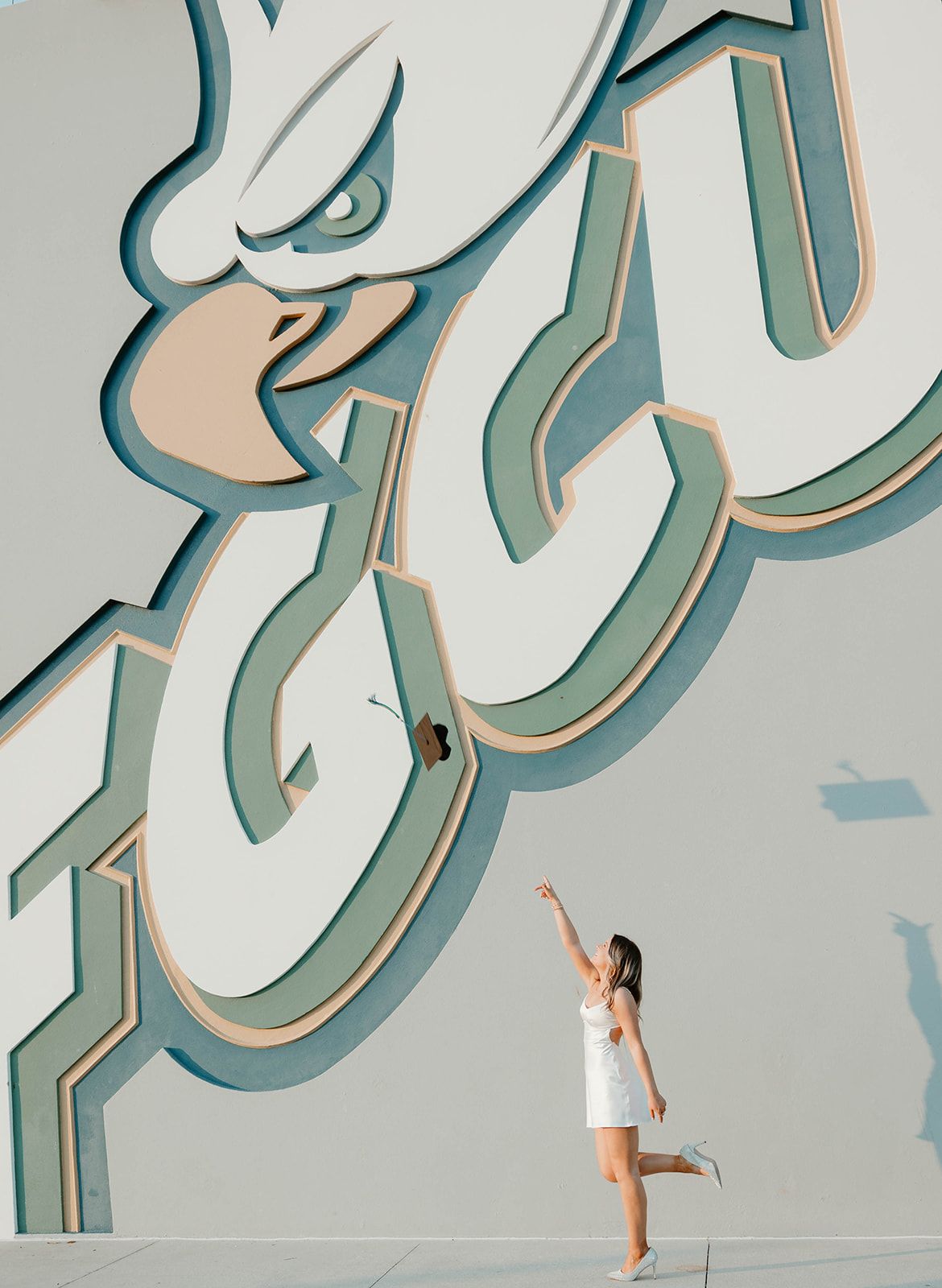 Person in white dress poses gracefully against geometric wall art installation with teal-colored 3D Arabic calligraphy.