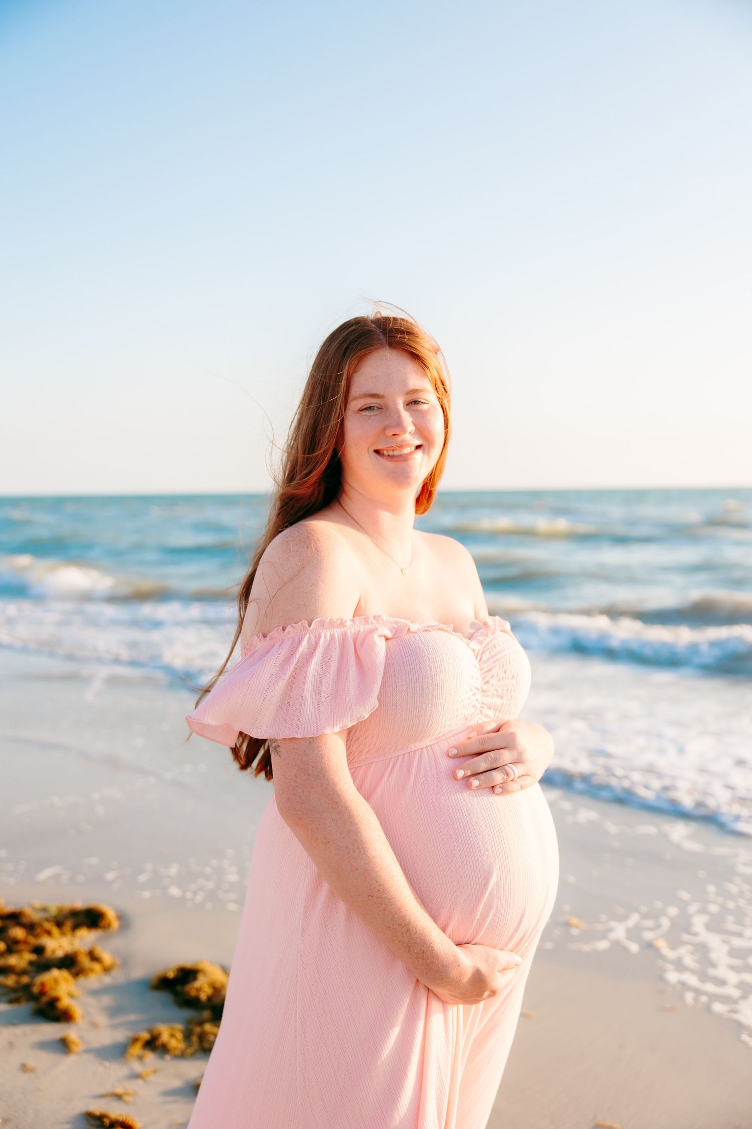 Smiling pregnant woman in pink off-shoulder dress cradling her baby bump on a sunny beach.