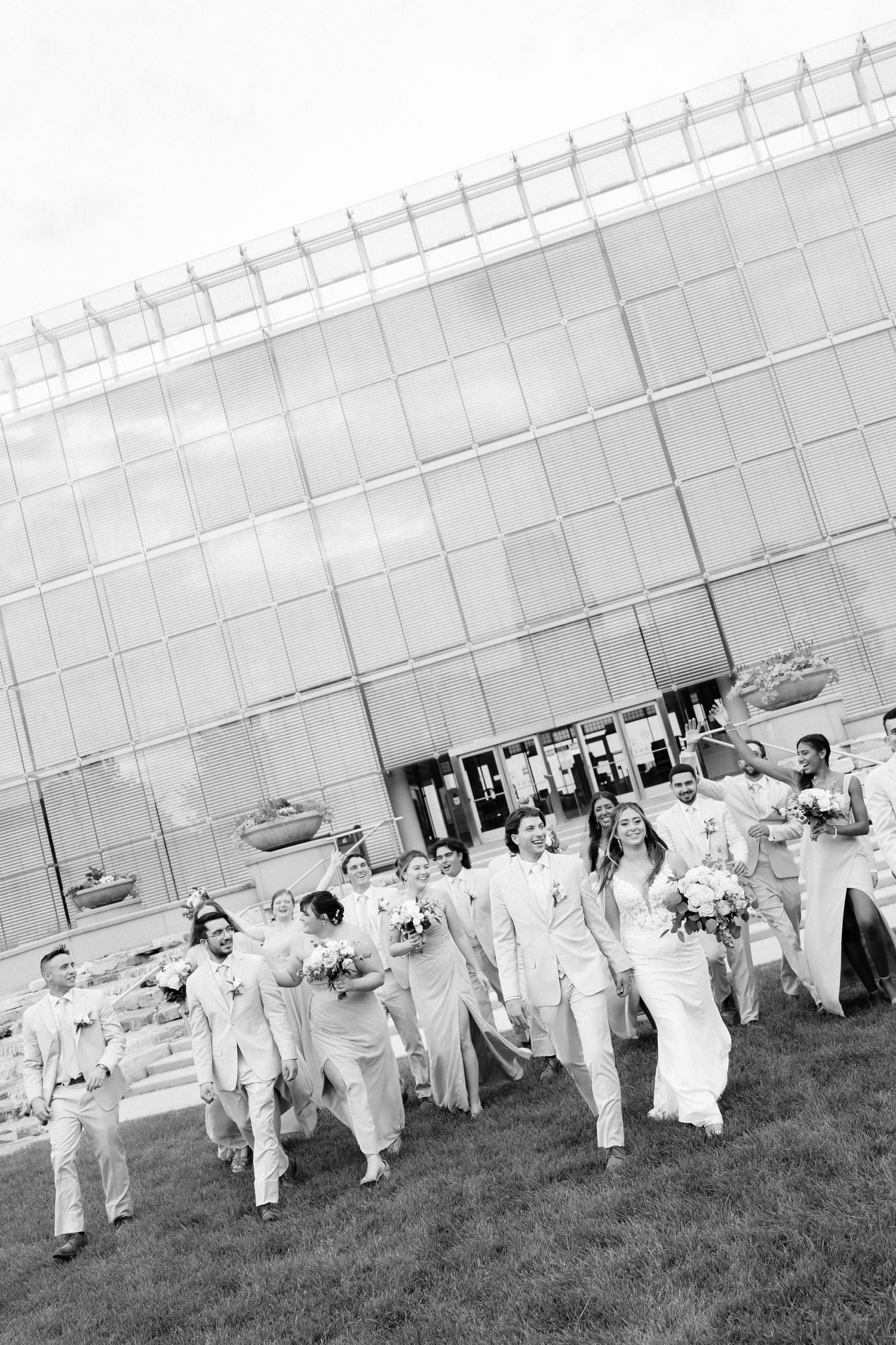 Black and white photo of a wedding party in white attire walking outdoors in front of a modern glass building.